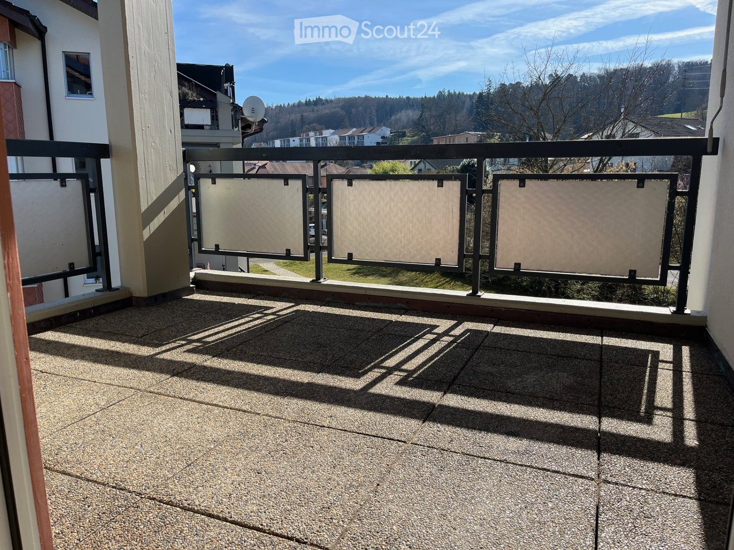 Balcony with a tiled concrete floor, black metal railing, and a view of a residential area with houses and trees.