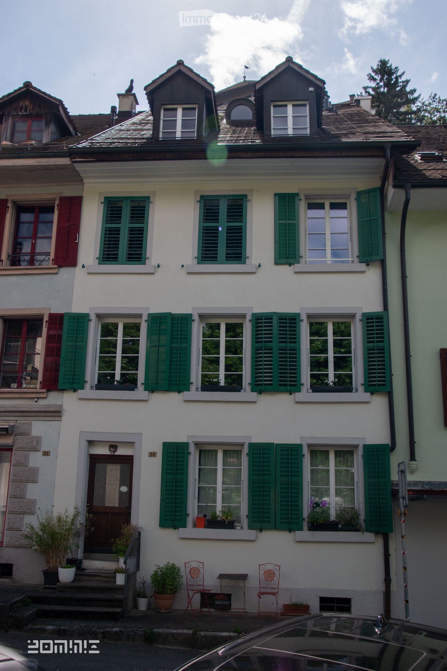 Three-story residential house with white facade, green shutters, multiple windows, and small balcony. Features potted plants and outdoor seating area with chairs and table.