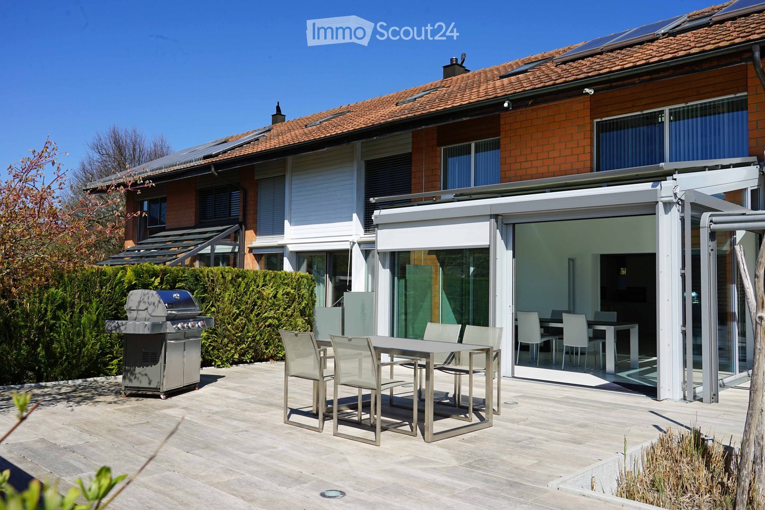 2-story brick house with a tiled roof, large windows, and a balcony. There is a patio area with outdoor furniture and a barbecue grill, as well as a garage and parking space on the property.