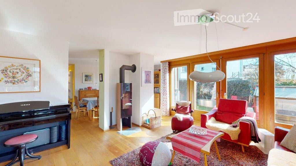 Bright living room with wooden flooring, large windows, red armchairs, a sofa, coffee table, piano, and a fireplace. The space is well-lit with natural light and features decorative elements like artwork and a woven basket.