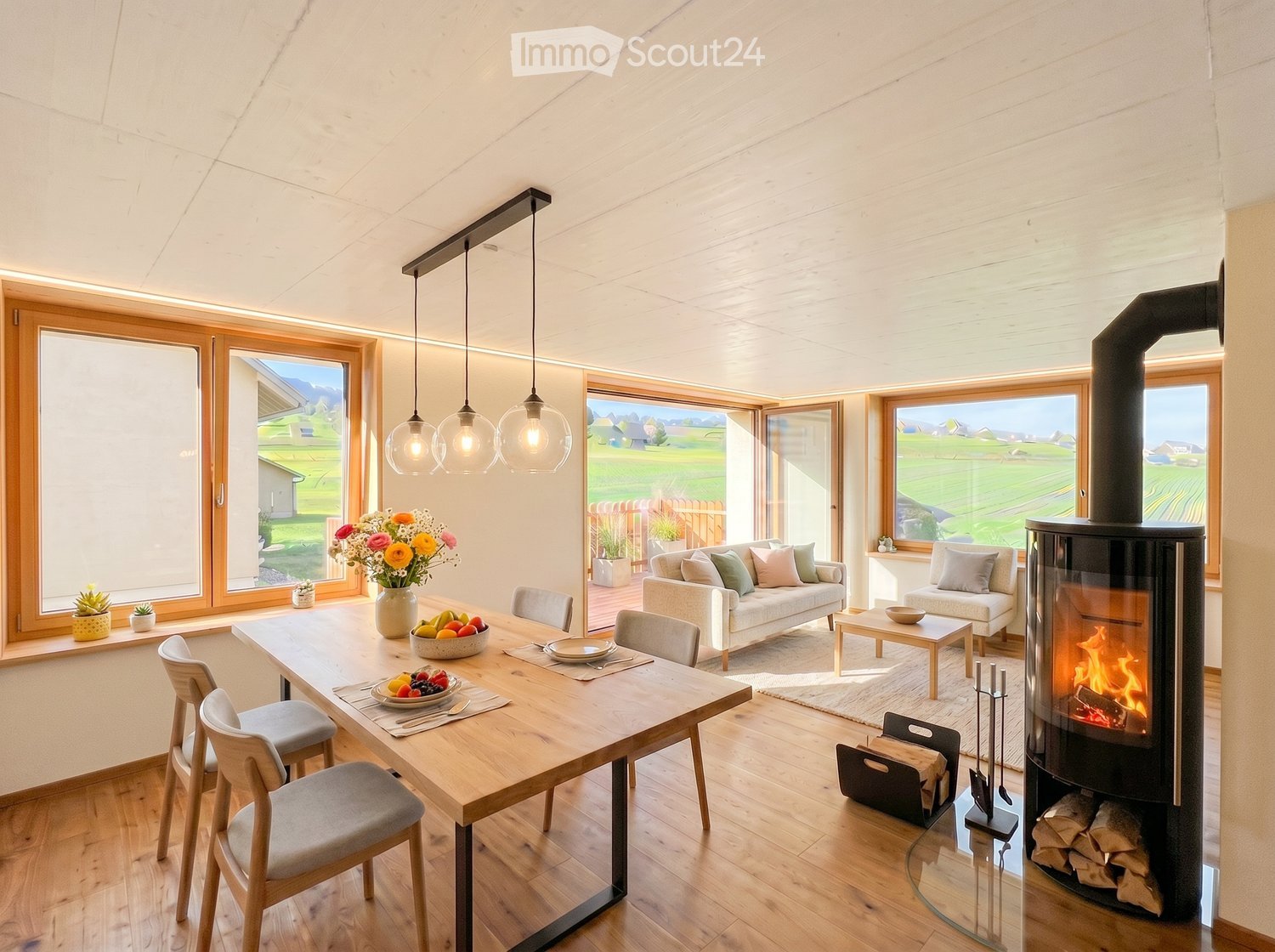 Bright living and dining area with large windows overlooking a green landscape, featuring a sofa, coffee table, dining table with chairs, and a modern wood-burning stove.