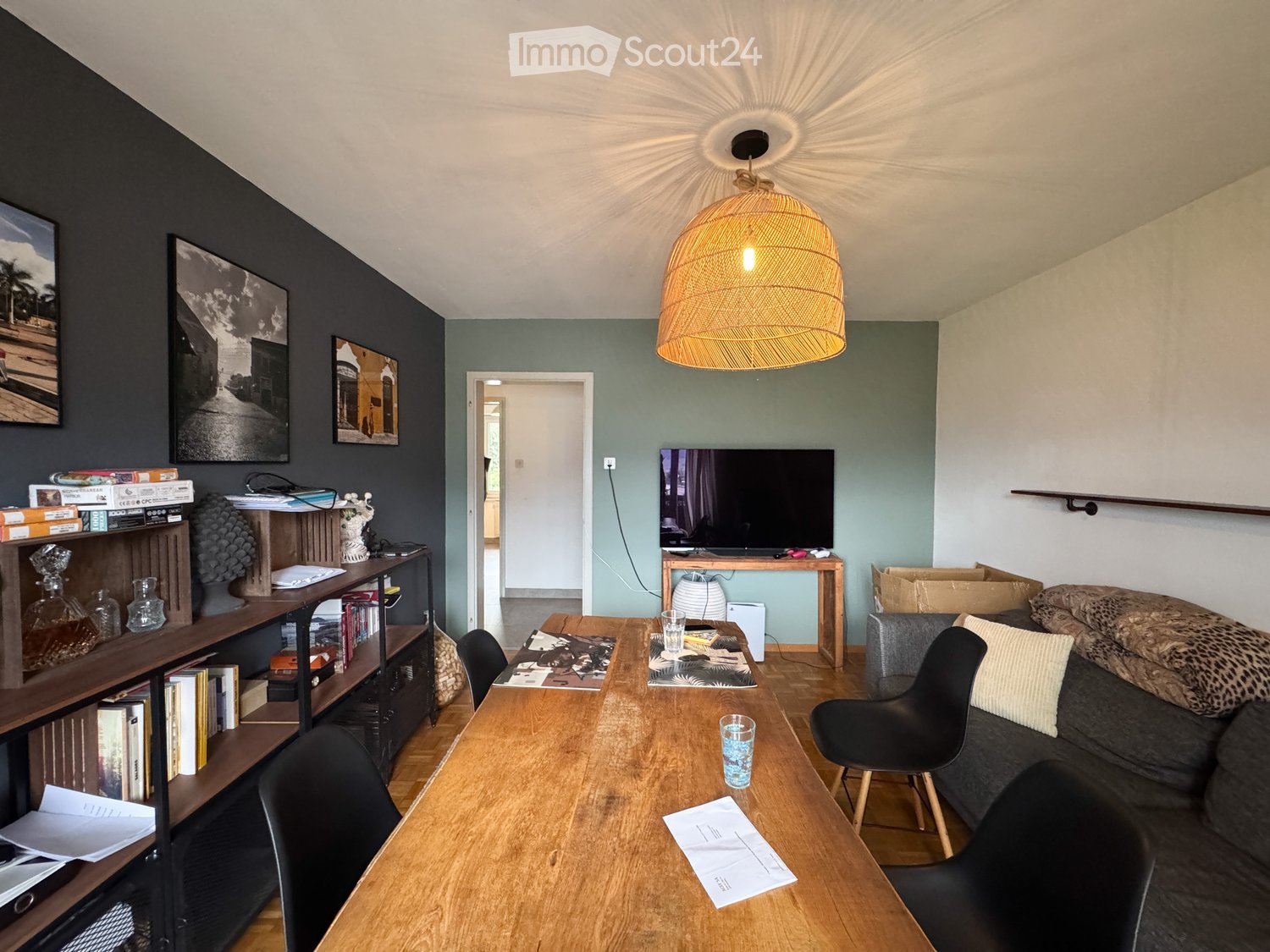 A modern living room with a dark grey sofa, wooden coffee table, black chairs, a shelf with books and decorative items, a television on a stand, and a hanging woven lamp.