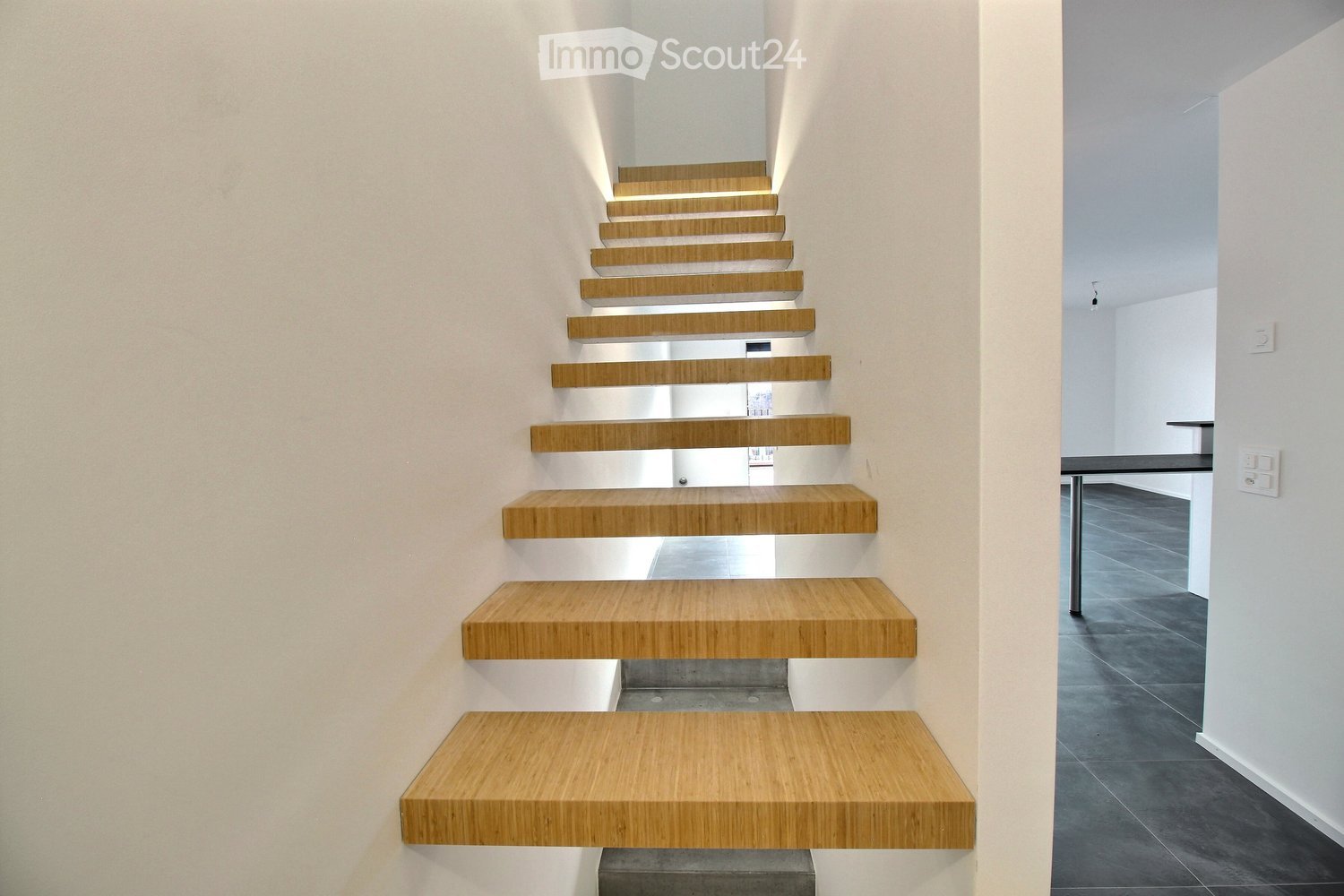 Modern staircase with wooden steps and white walls, leading to an open area with dark flooring and minimal furnishings.