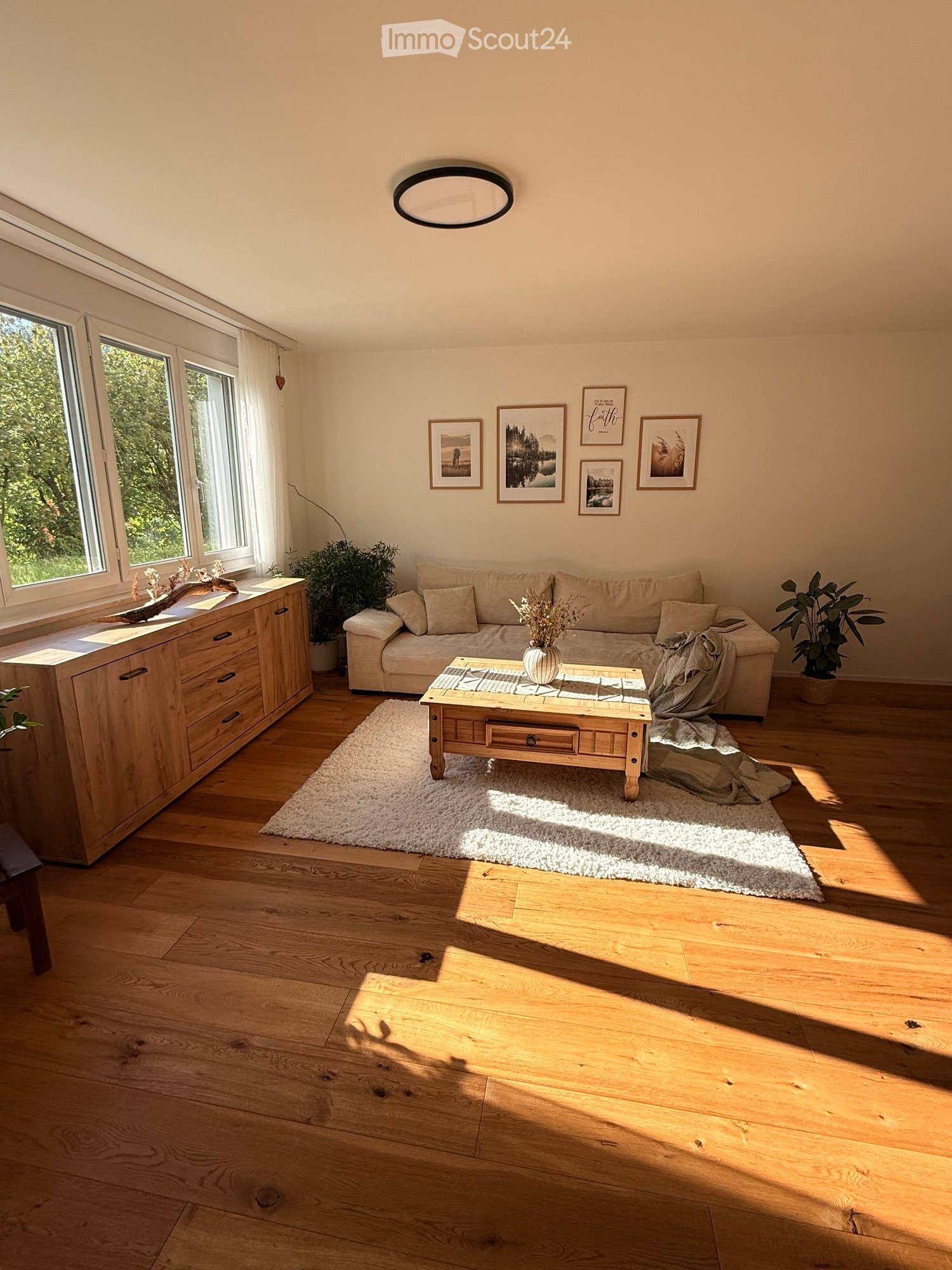Bright living room with wooden flooring, large windows, a cream-colored sofa, a wooden coffee table with a vase, a sideboard, and several picture frames on the wall.