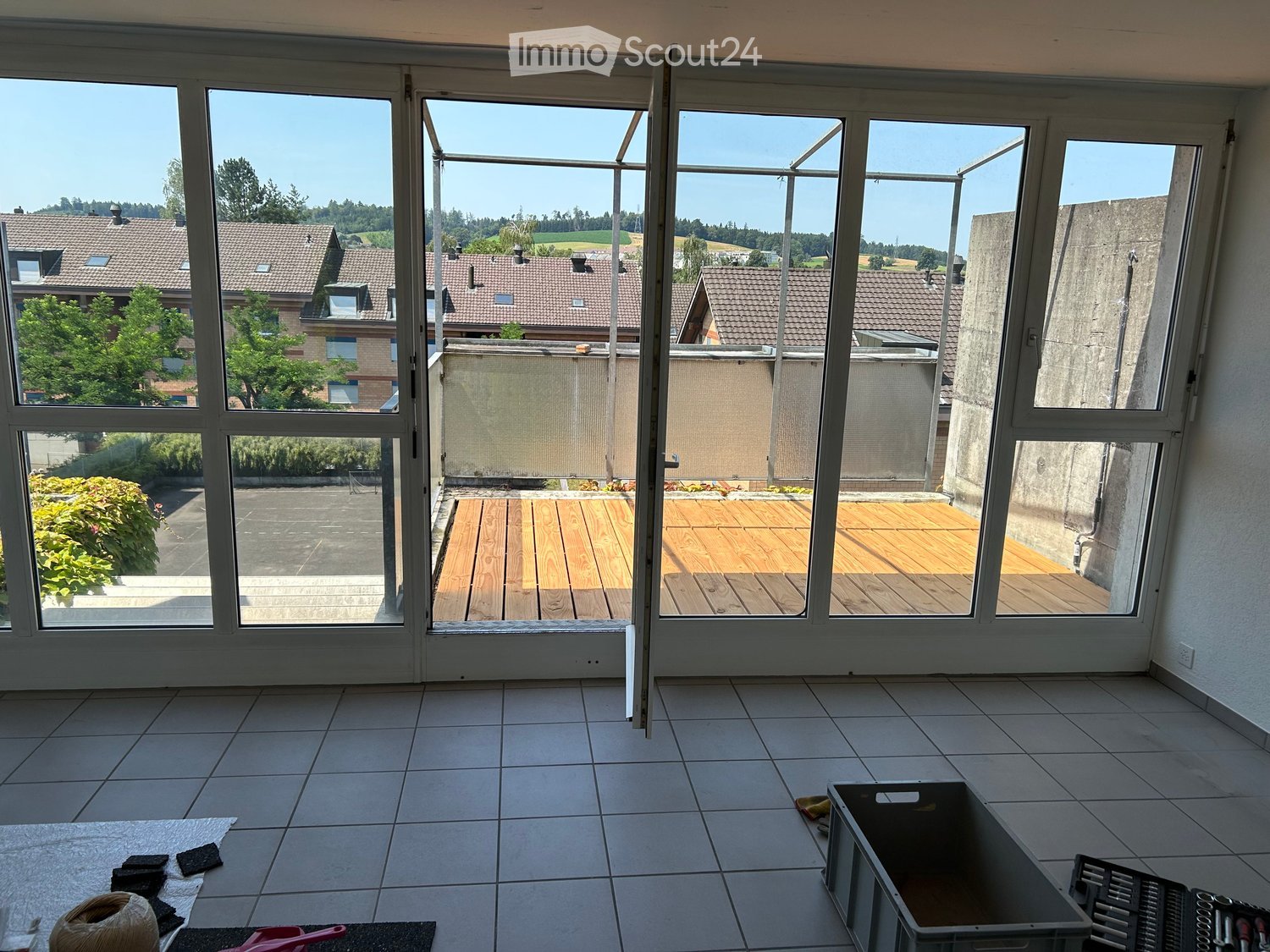 Empty room with tiled floor, large glass doors leading to a wooden deck and outdoor area, visible tools and materials on the floor.