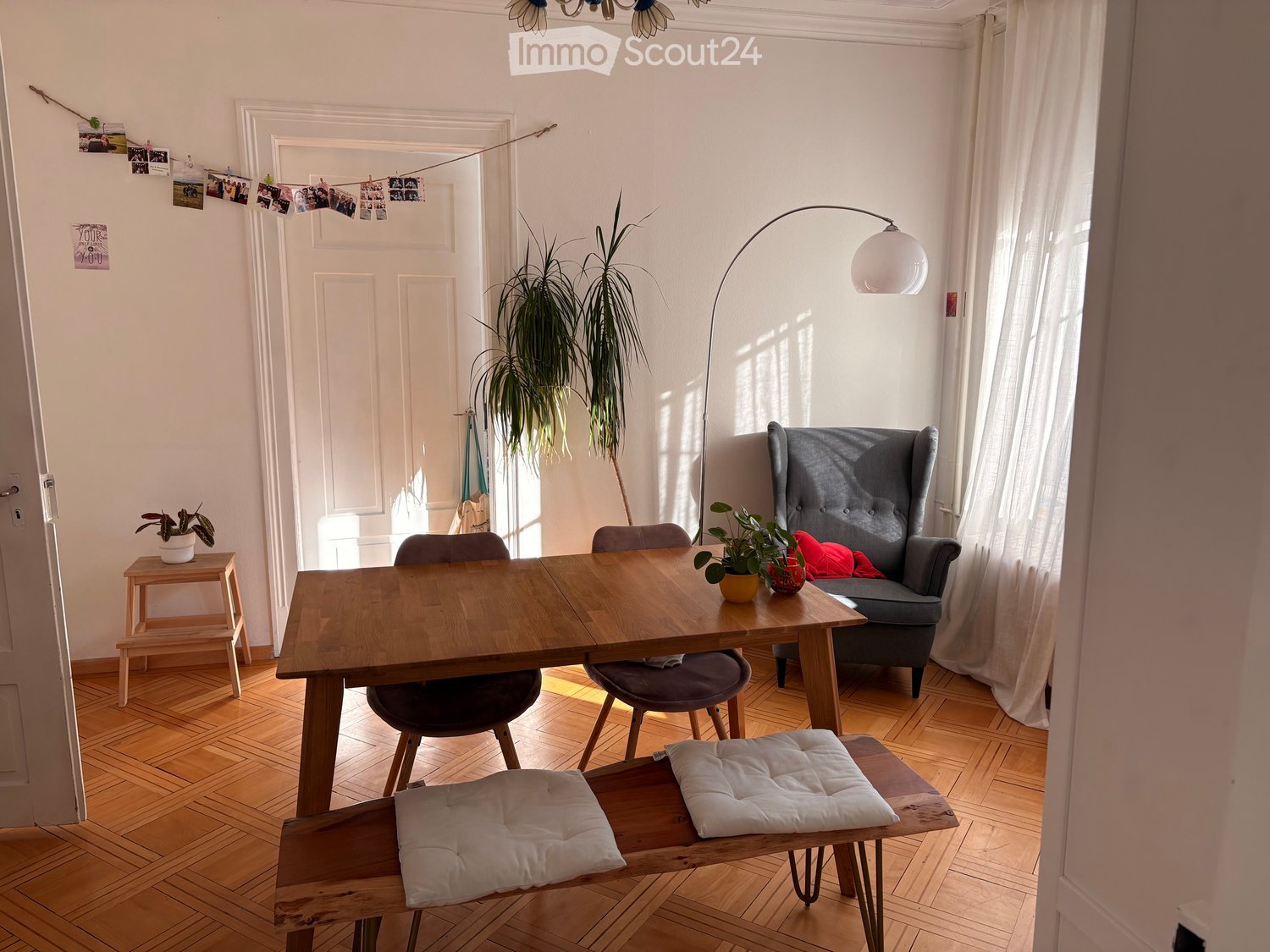 Bright living room with wooden flooring, large window with white curtains, grey sofa, wooden dining table with chairs, floor lamp, and potted plants.