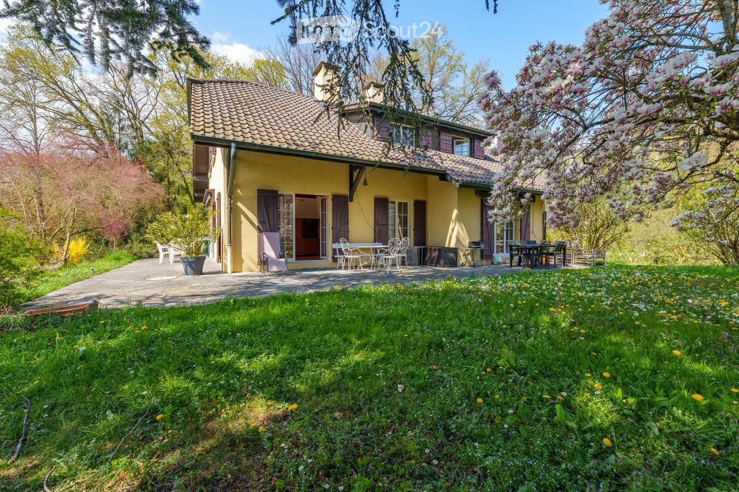 A two-story yellow house with a tiled roof, surrounded by a well-maintained garden with a patio area, outdoor furniture, and blooming trees.