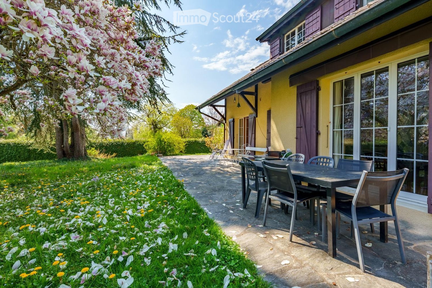 A well-maintained garden area with a patio seating arrangement, including a table and chairs, surrounded by blooming flowers and trees. The house in the background has a yellow facade and large windows.