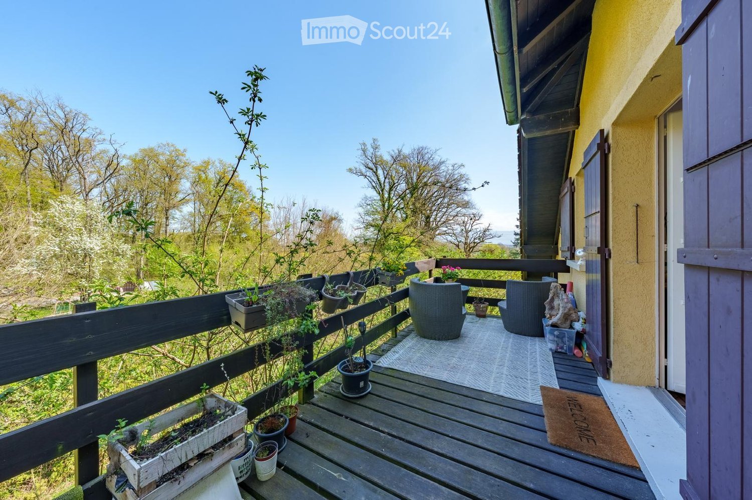 Wooden balcony with black railing, several potted plants, two chairs, and a welcome mat.