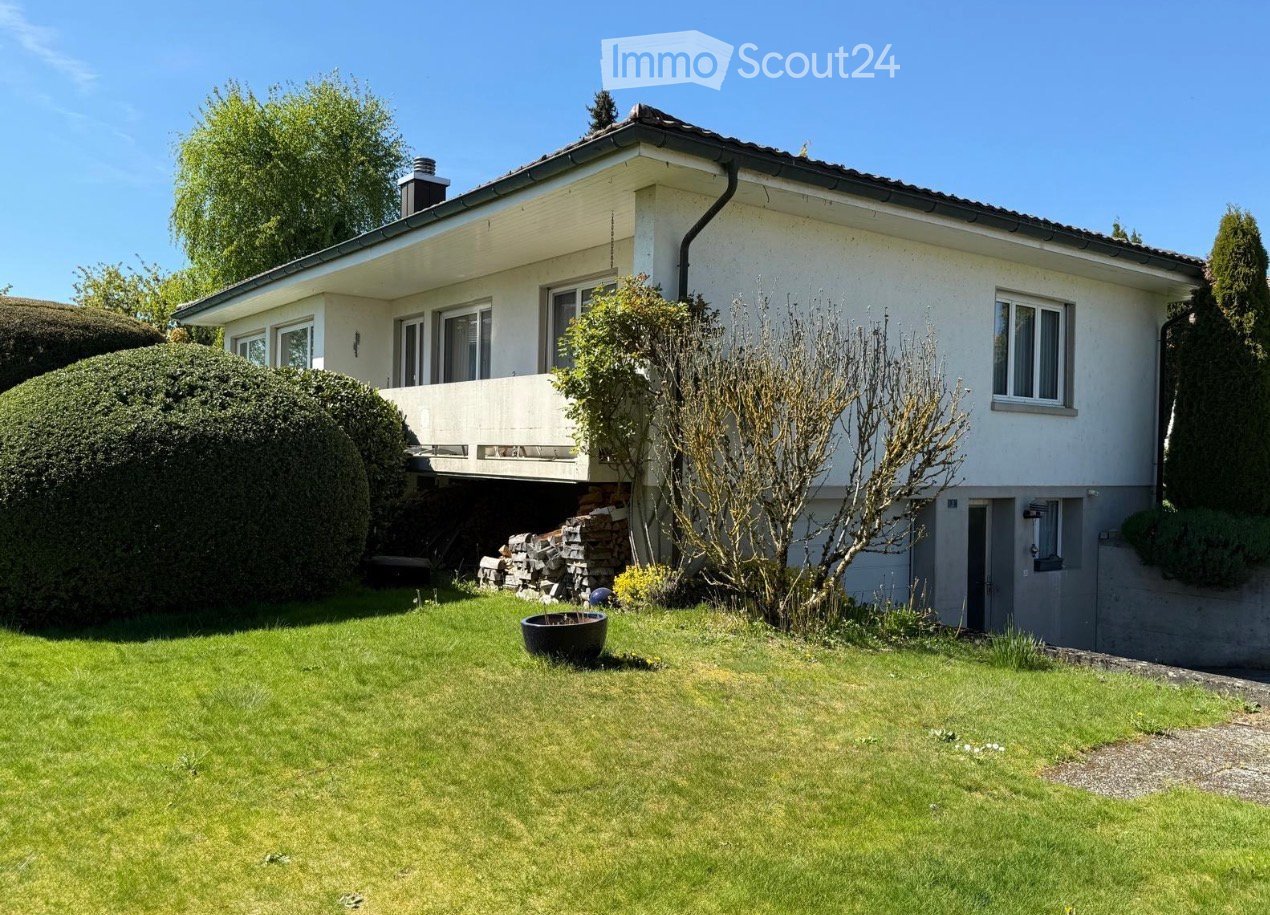 Two-story house with white exterior, multiple windows, and a balcony. The house is surrounded by a well-maintained lawn, shrubs, and trees.