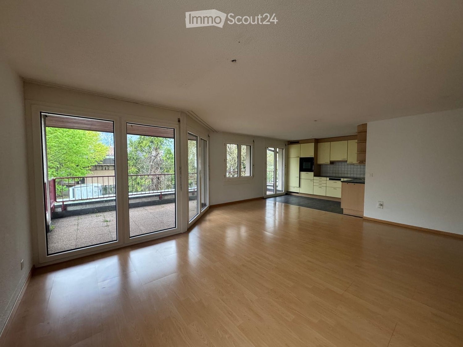 Empty living space with wooden flooring, large sliding glass doors leading to a balcony, and a small kitchen area with cabinets and appliances visible in the background.