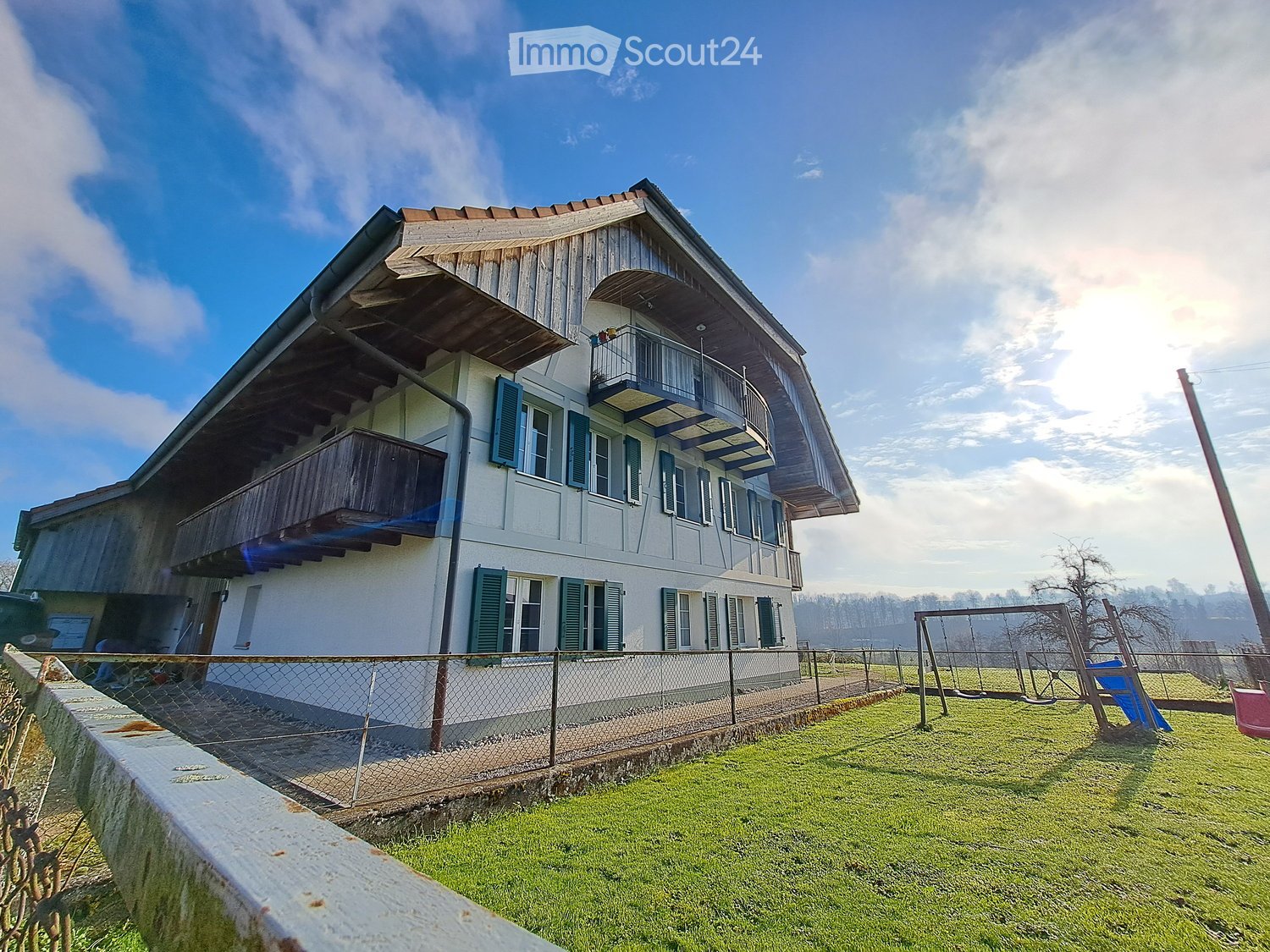 Two-story house with white walls, green shutters, and wooden balconies. The house is surrounded by a fenced yard with a swing set and grassy area.