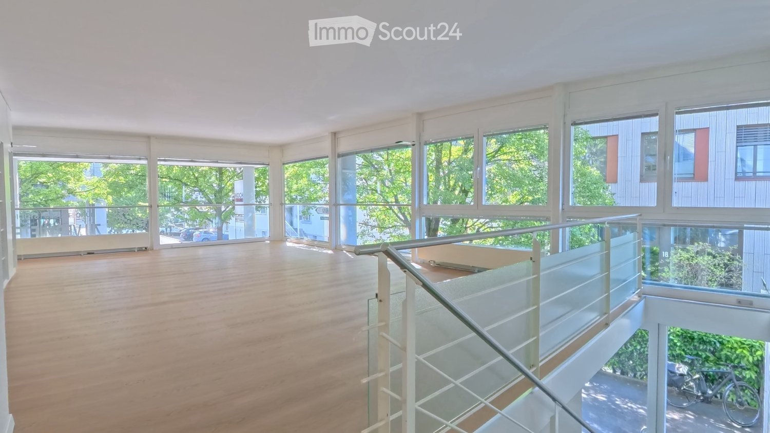 Open, empty room with wooden flooring and large windows, featuring a modern glass staircase leading to an upper level.
