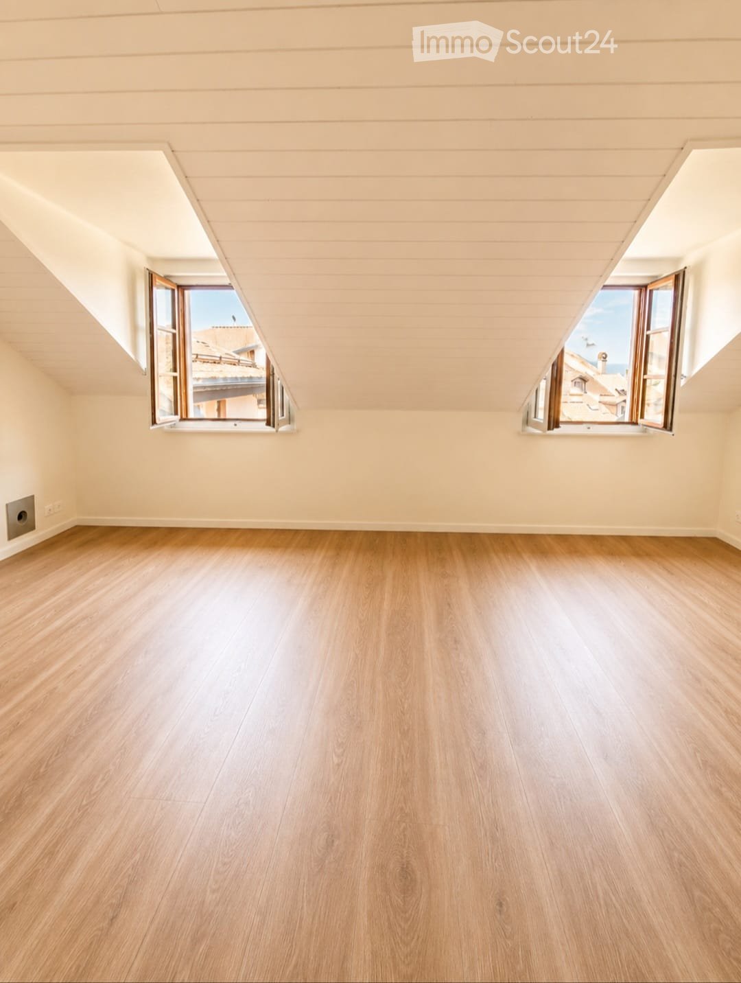 Empty attic room with wooden flooring, slanted ceilings, and two large windows offering a view of the outside.