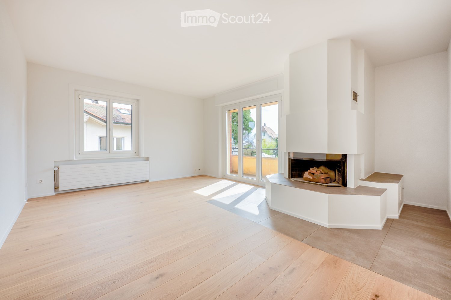 Empty living room with light wooden flooring, white walls, large windows, and a fireplace with a white surround.