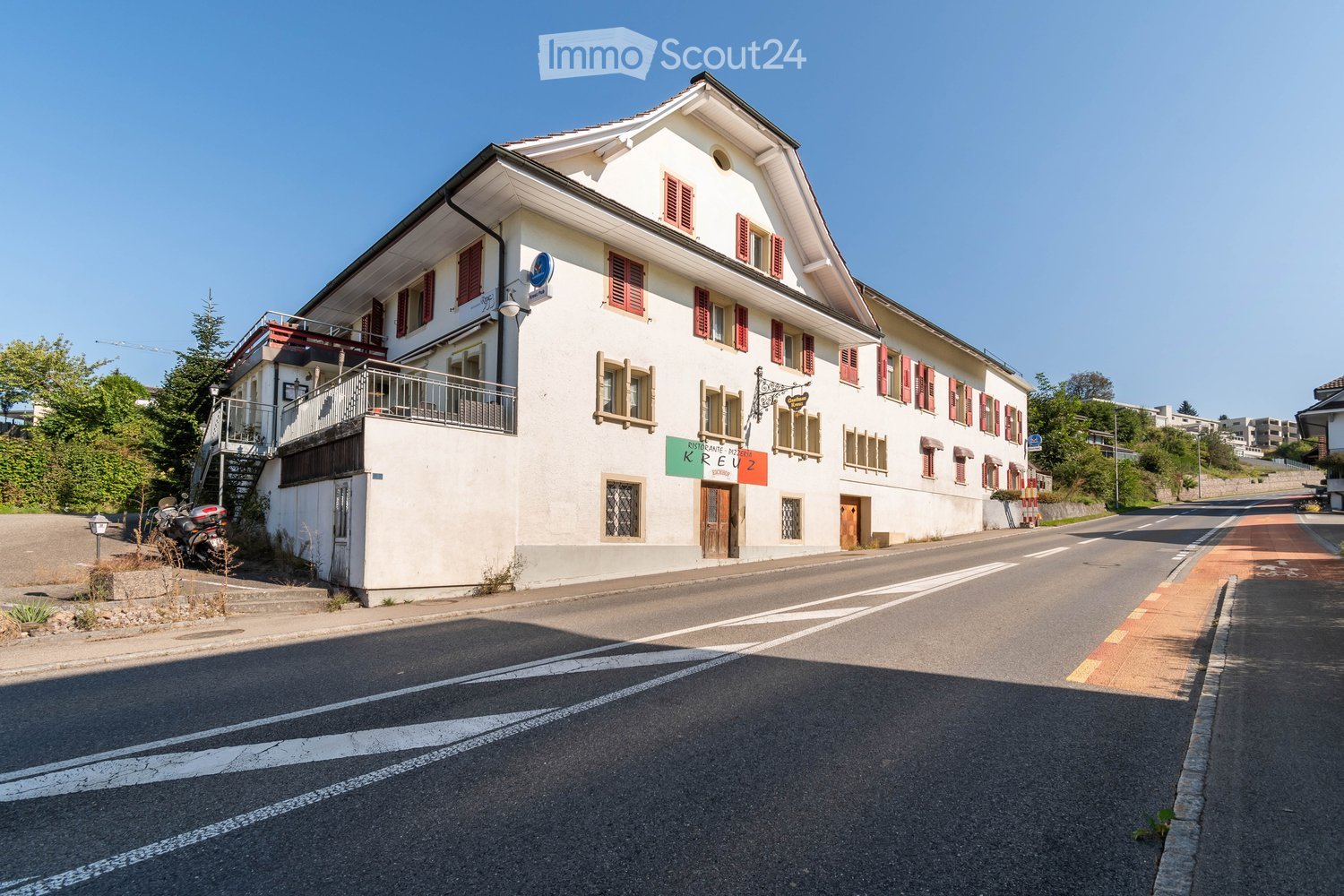 A two-story white building with red shutters and a balcony, featuring a sign for "Ristorante Pizzeria Kreuz" and a motorcycle parked nearby.