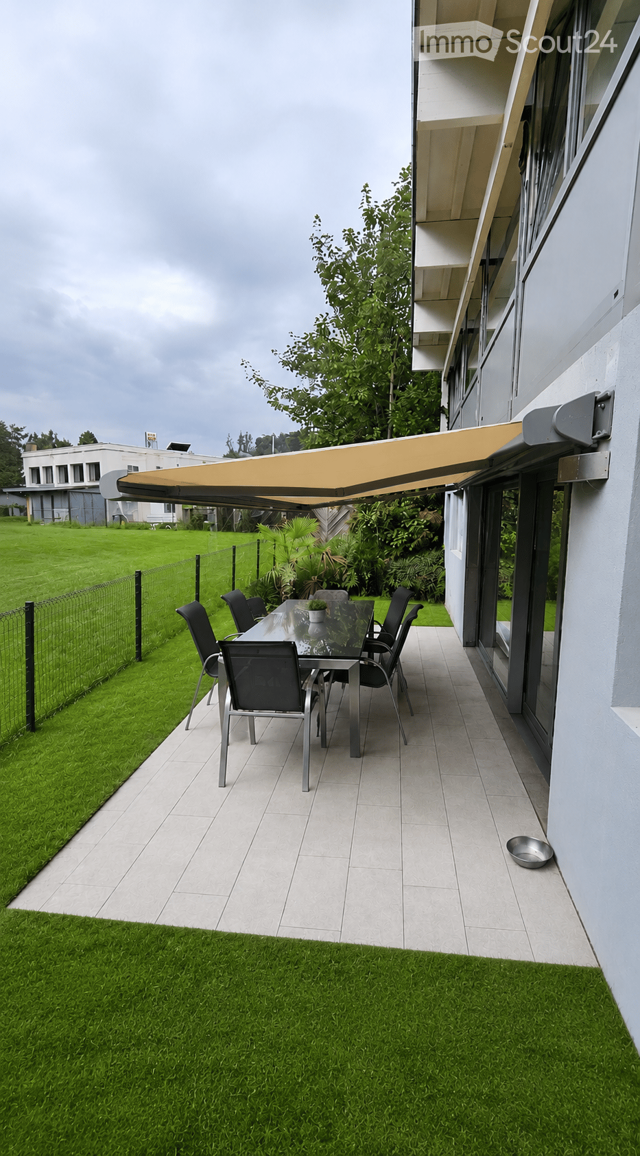 Outdoor patio with a glass dining table, six chairs, and a small potted plant, covered by a retractable awning attached to a modern building.