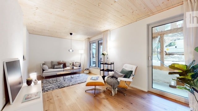 Modern living room with light wooden flooring, white walls, and a wooden ceiling. Features a grey sofa with throw pillows, a coffee table, an armchair with an ottoman, a floor lamp, a television, and a side table with a vase.