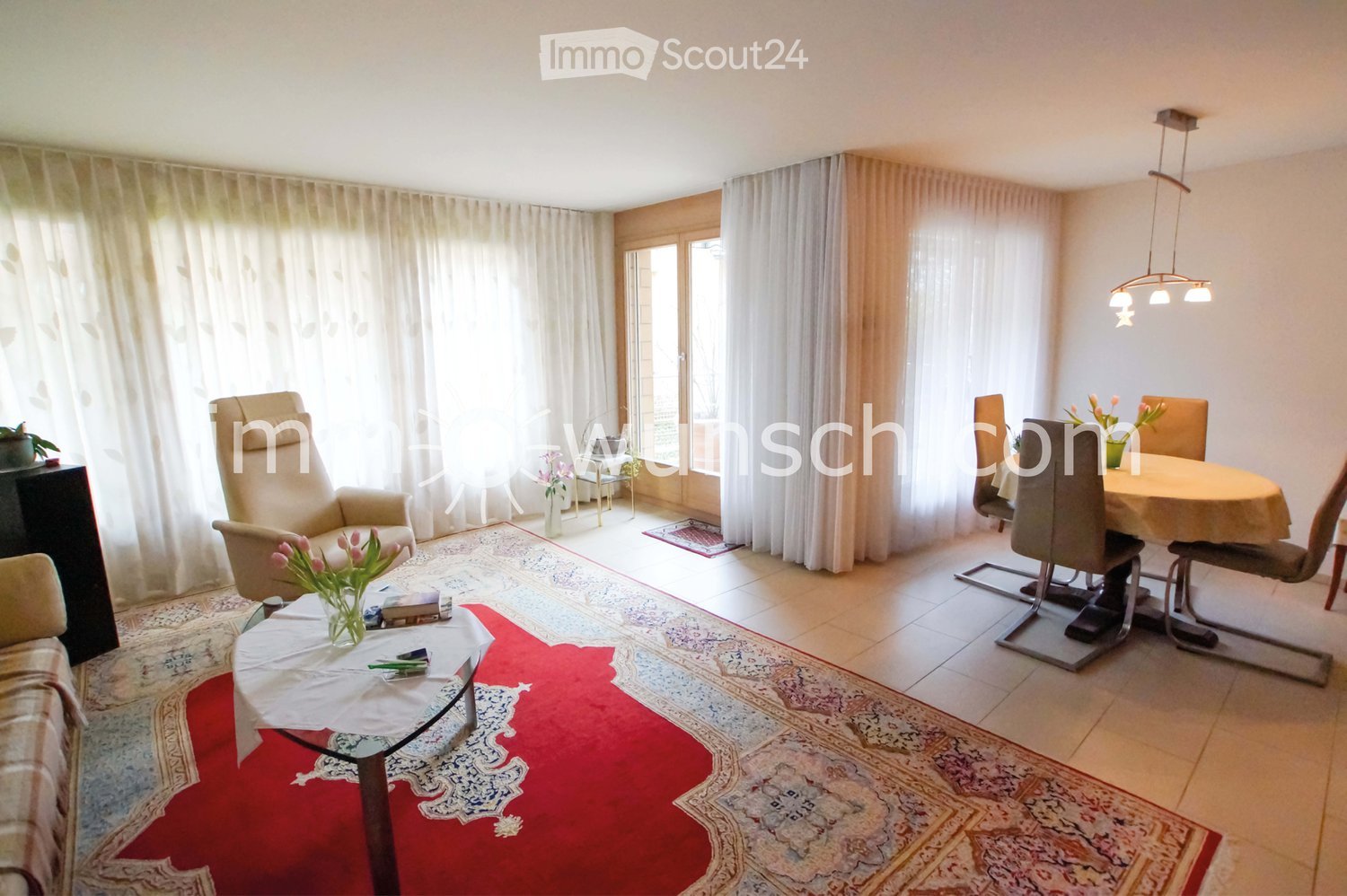 Bright living and dining area with large windows, beige curtains, patterned rugs, a sofa, coffee table, dining table with chairs, and decorative vases.