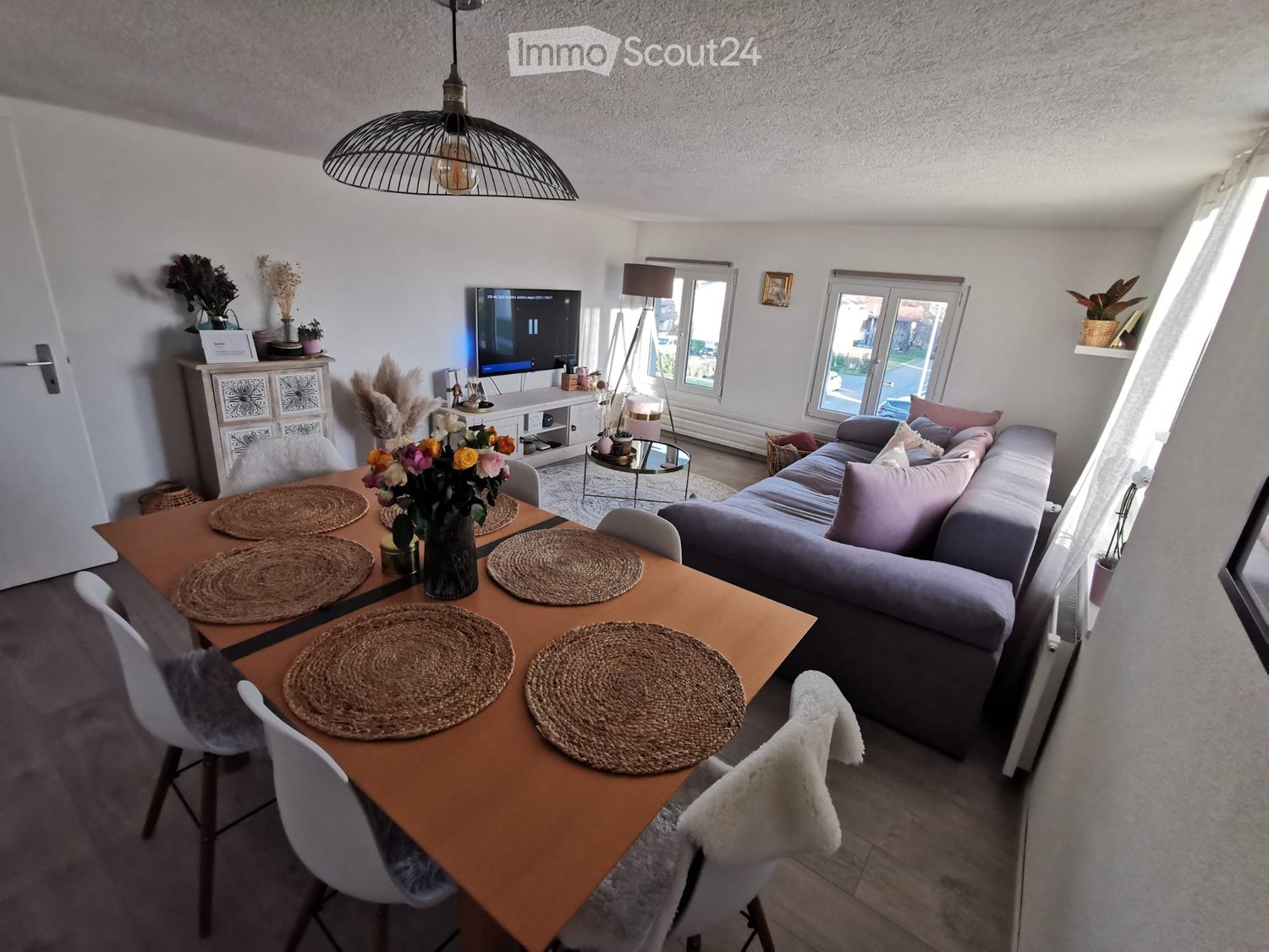 Modern living and dining area with a grey sofa, wooden dining table with six chairs, a television, and decorative items including a flower vase and a floor lamp.