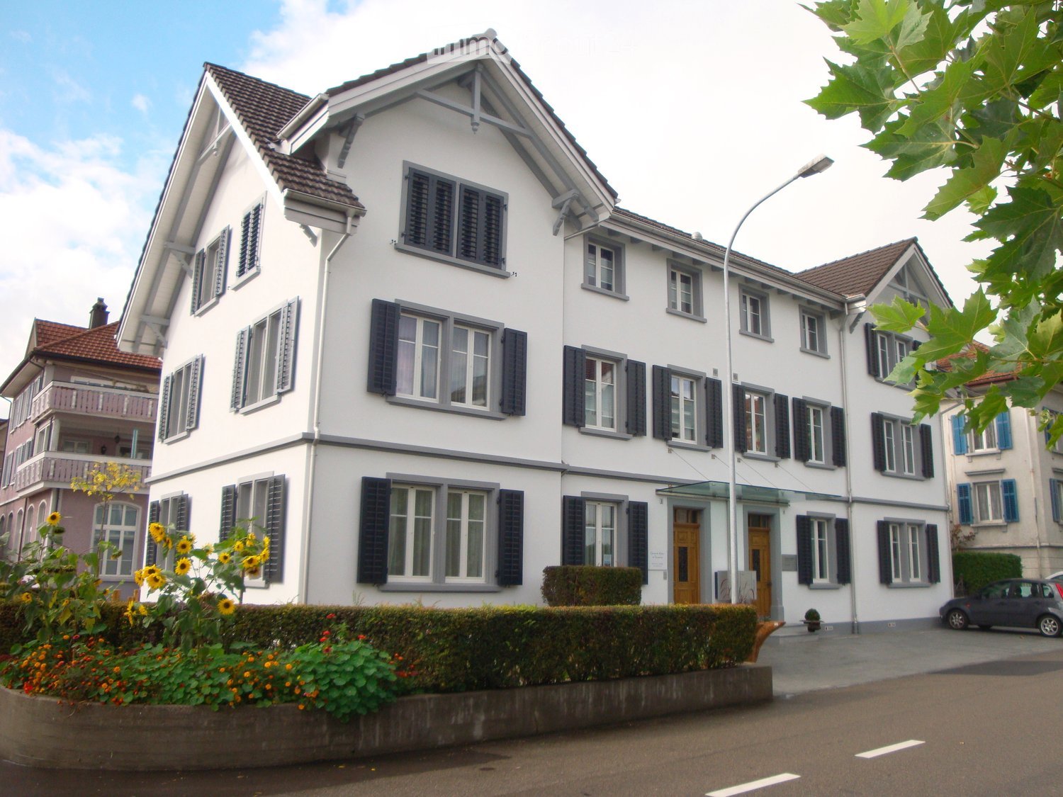 A multi-story white house with dark shutters, multiple windows, and a well-maintained garden in front.