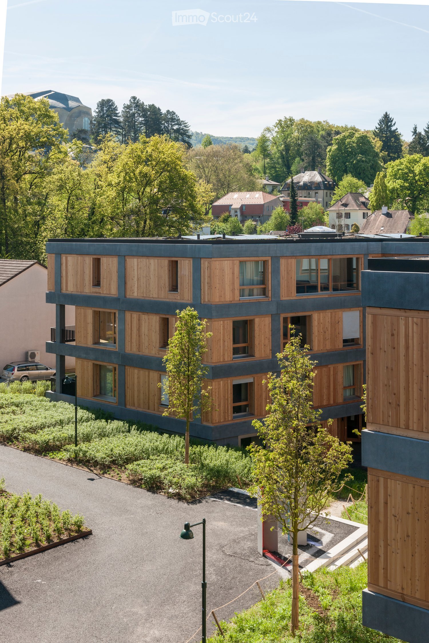 A modern multi-story residential building, wood cladding, surrounded by greenery, several windows