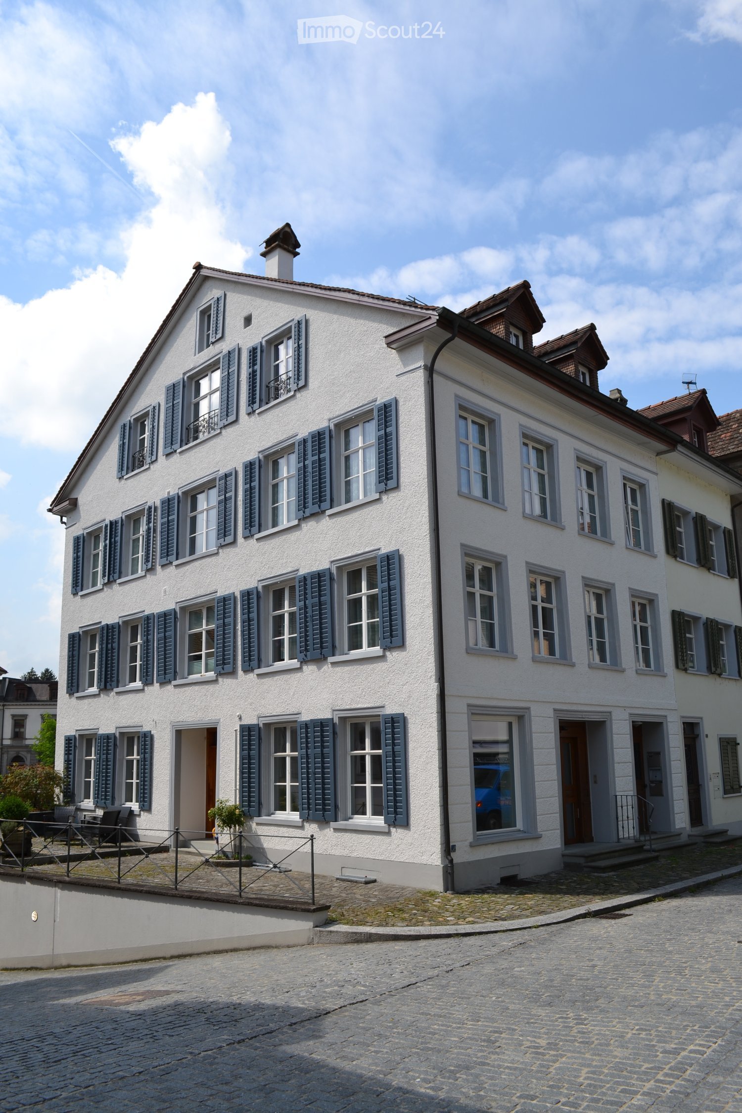 white building, multiple windows, blue shutters, brown roof, paved street, small garden