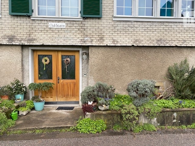 Front view of a two-story house with a wooden double door, green shutters, and a small garden with potted plants and shrubs.