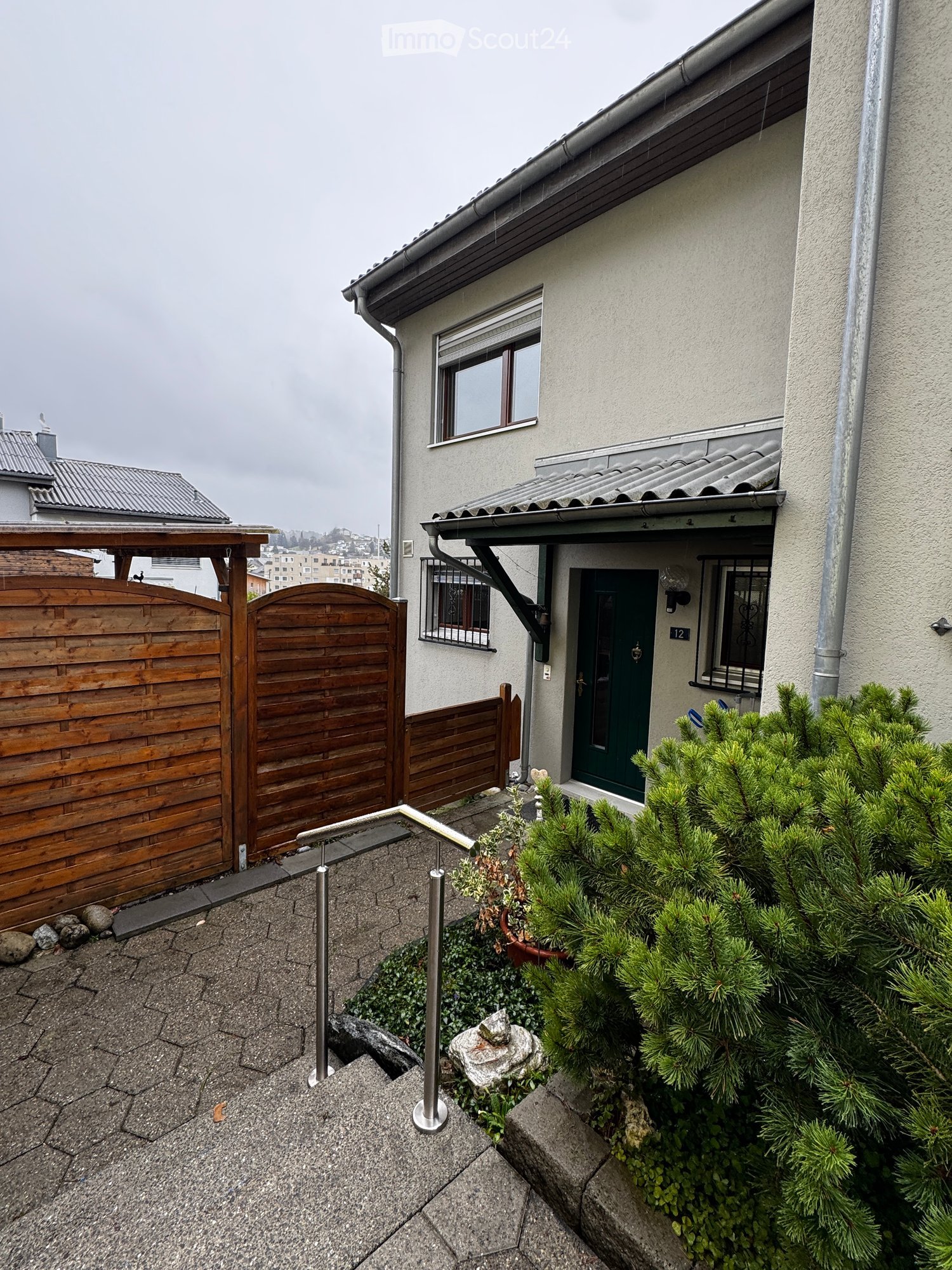 Two-story house with a green front door, wooden gate, and small garden area with plants and a stone sculpture.