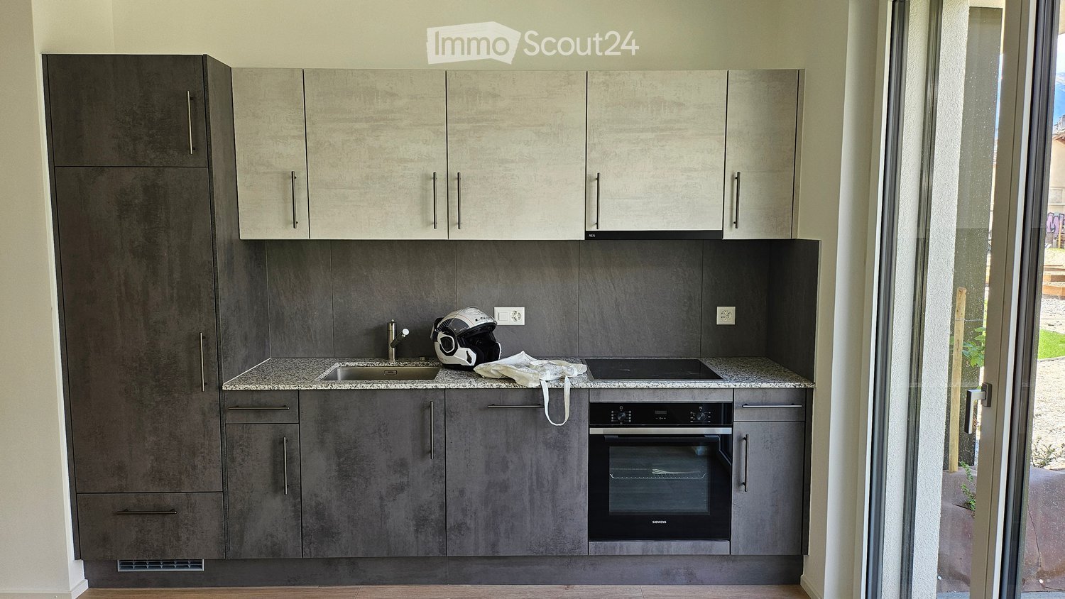 Modern kitchen with grey cabinets, granite countertops, stainless steel sink, and built-in oven. A helmet and cloth are placed on the counter.