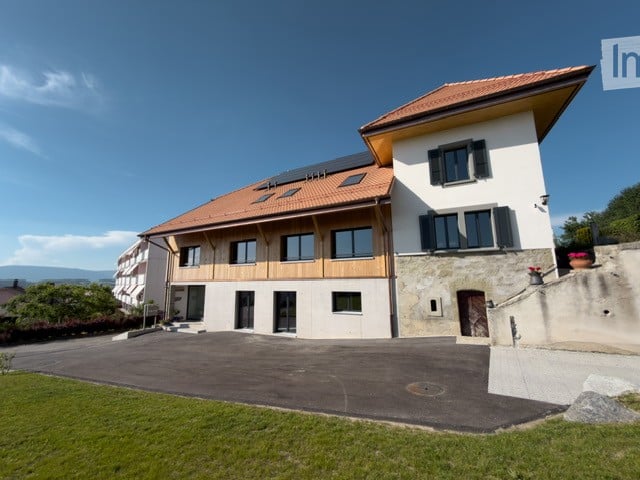 Modern two-story house with white walls, dark wooden accents, and a red-tiled roof. Features large windows, a driveway, and a well-maintained lawn.