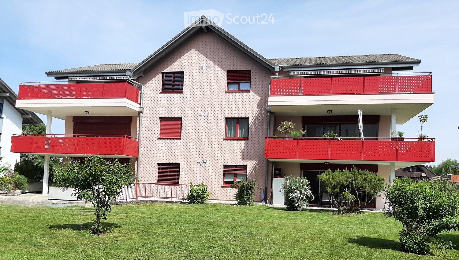 Two-story residential building with pink facade, red balconies, multiple windows, and a well-maintained lawn.