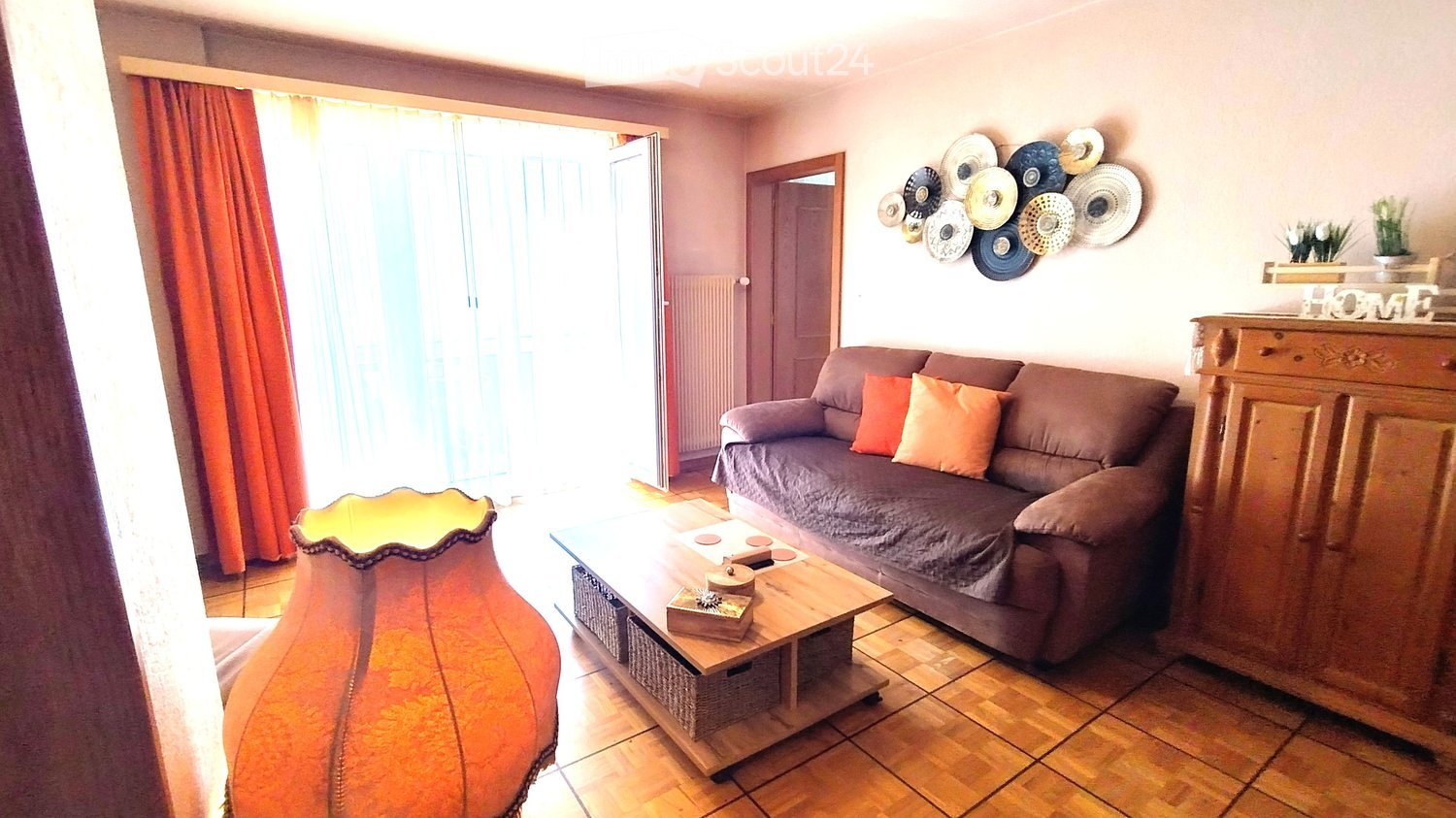 Living room with a grey sofa, orange cushions, a wooden coffee table, a floor lamp, and decorative plates on the wall.