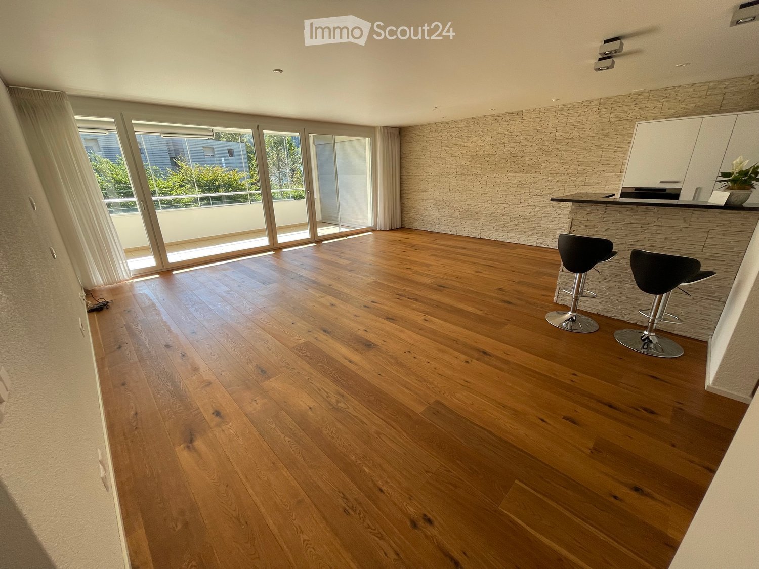 Open-plan kitchen and living area with wooden flooring, large windows leading to a balcony, and a kitchen island with two stools.