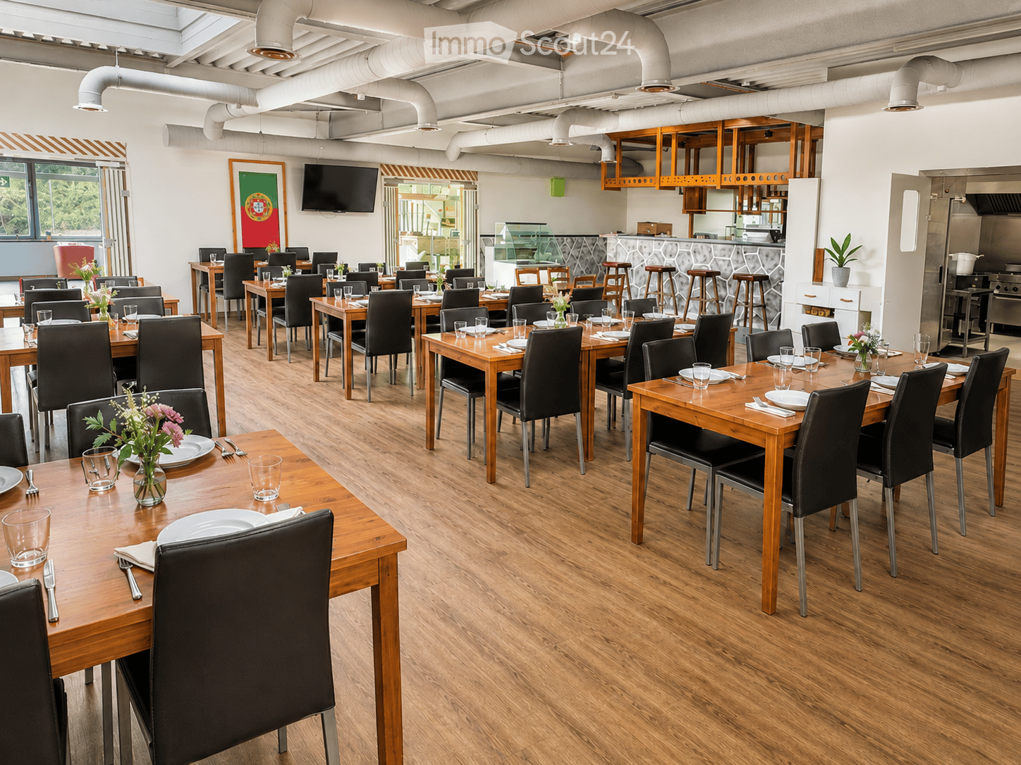 Restaurant interior with wooden tables, black chairs, and neatly set place settings, including plates, cutlery, and glassware. A bar area with stools and a television is visible in the background.