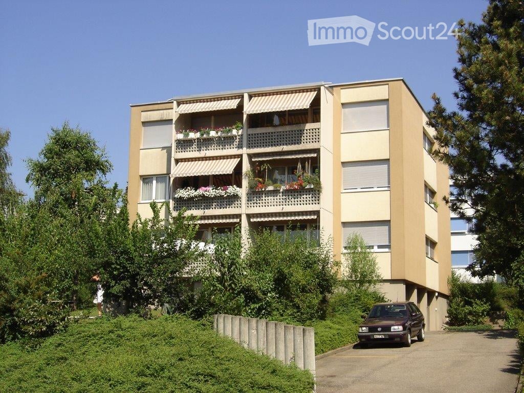 Four-story residential building with balconies, awnings, and flower boxes, surrounded by greenery and a parking area.