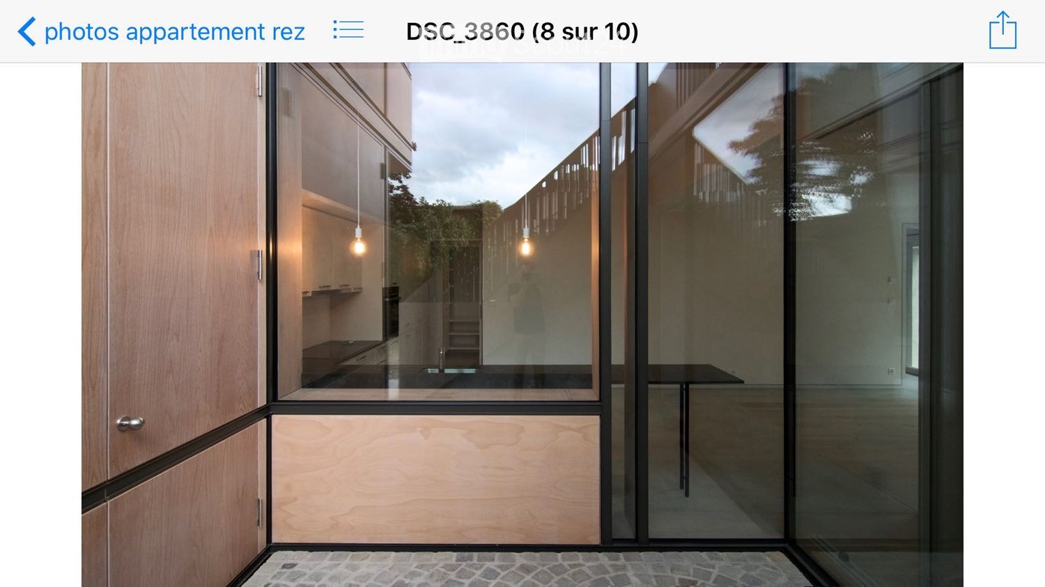 Modern hallway with large glass doors leading to a kitchen area, wooden cabinets, and pendant lights visible through the glass.
