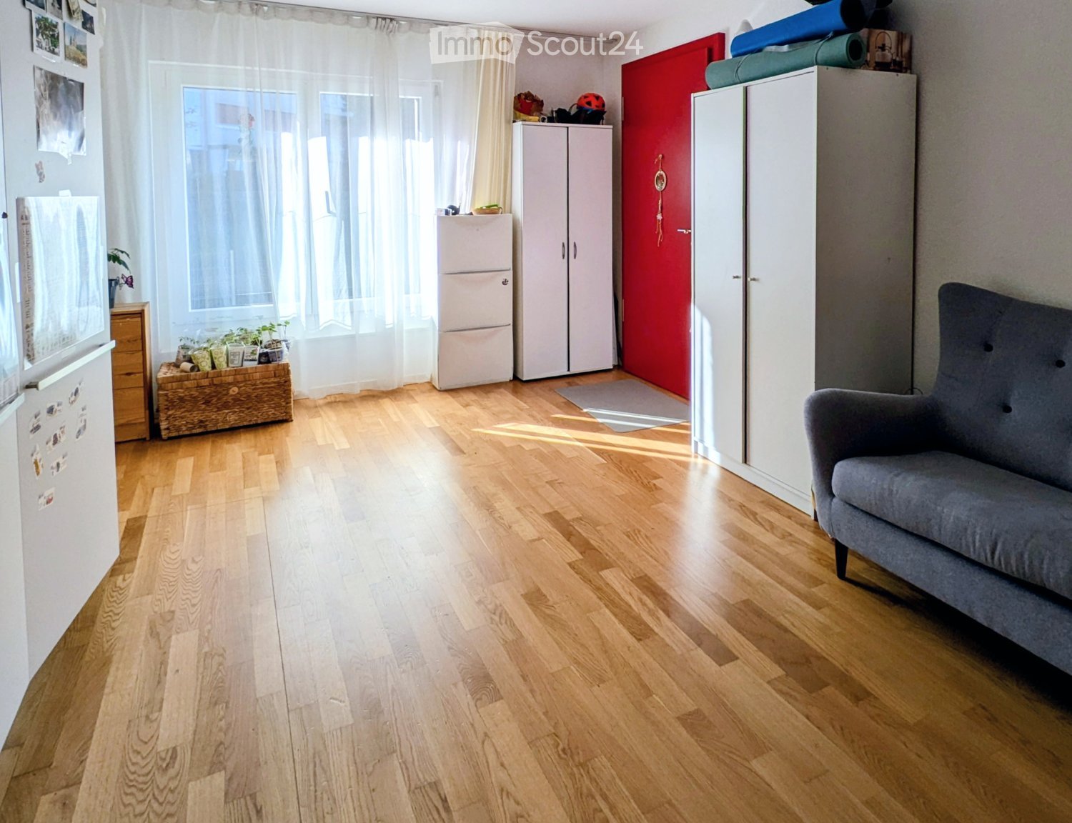 A well-lit room with wooden flooring, a grey sofa, a refrigerator, a cabinet, and a small plant on a wooden chest.
