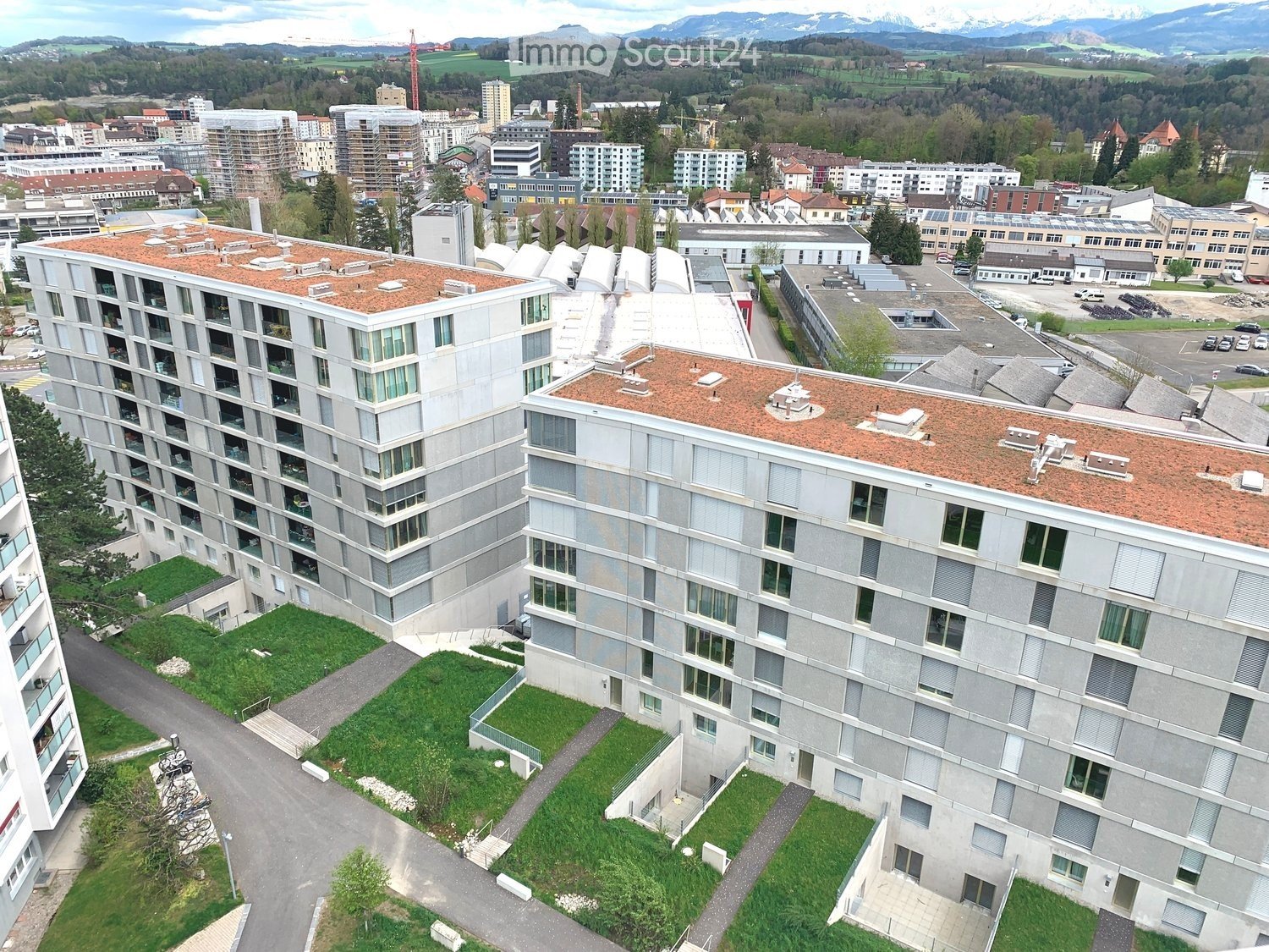 A modern multi-story residential building with a flat roof and numerous windows, surrounded by greenery and other structures in an urban setting.