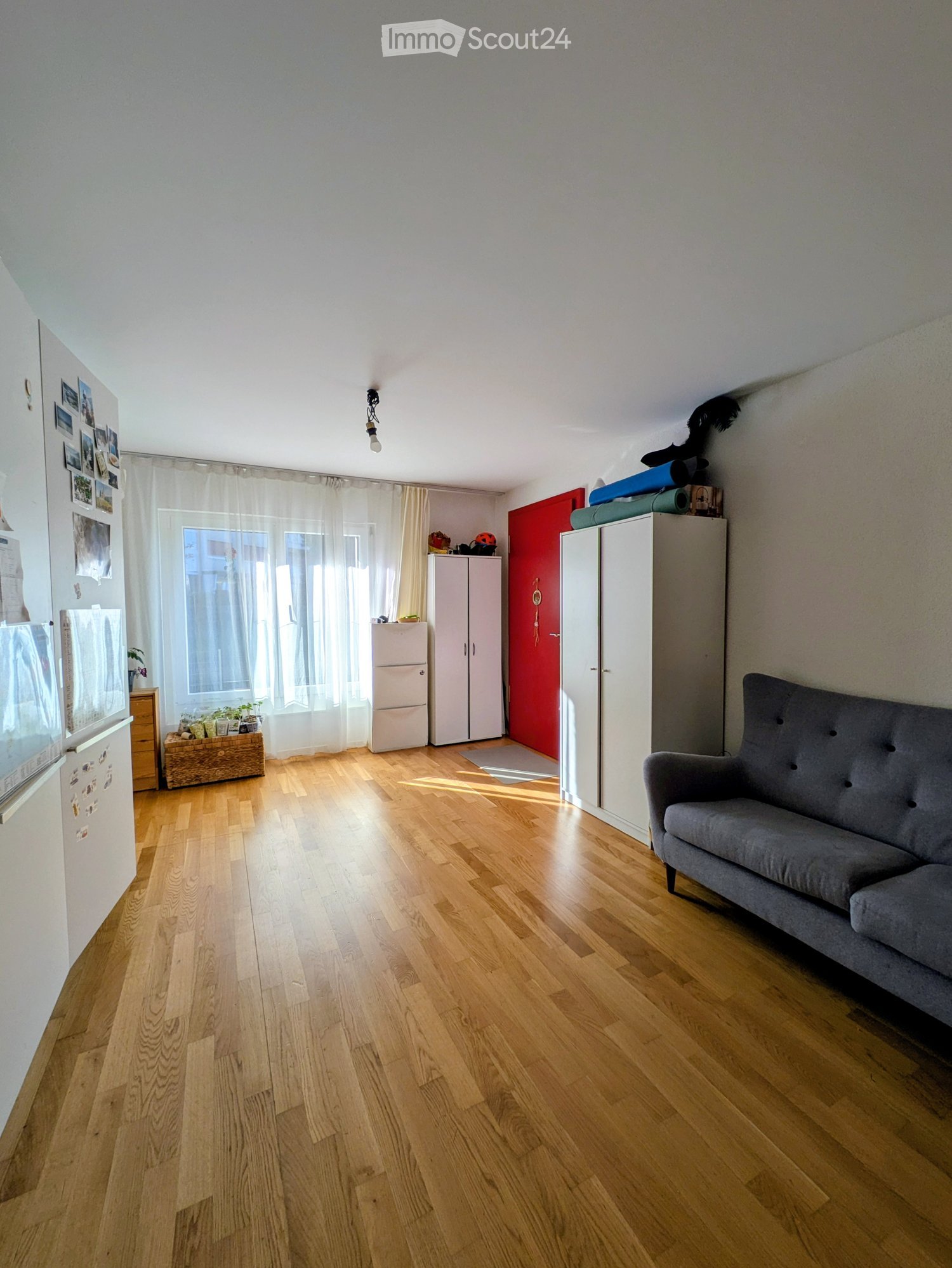 A well-lit living space with wooden flooring, a grey sofa, a refrigerator, cabinets, and a window with curtains.
