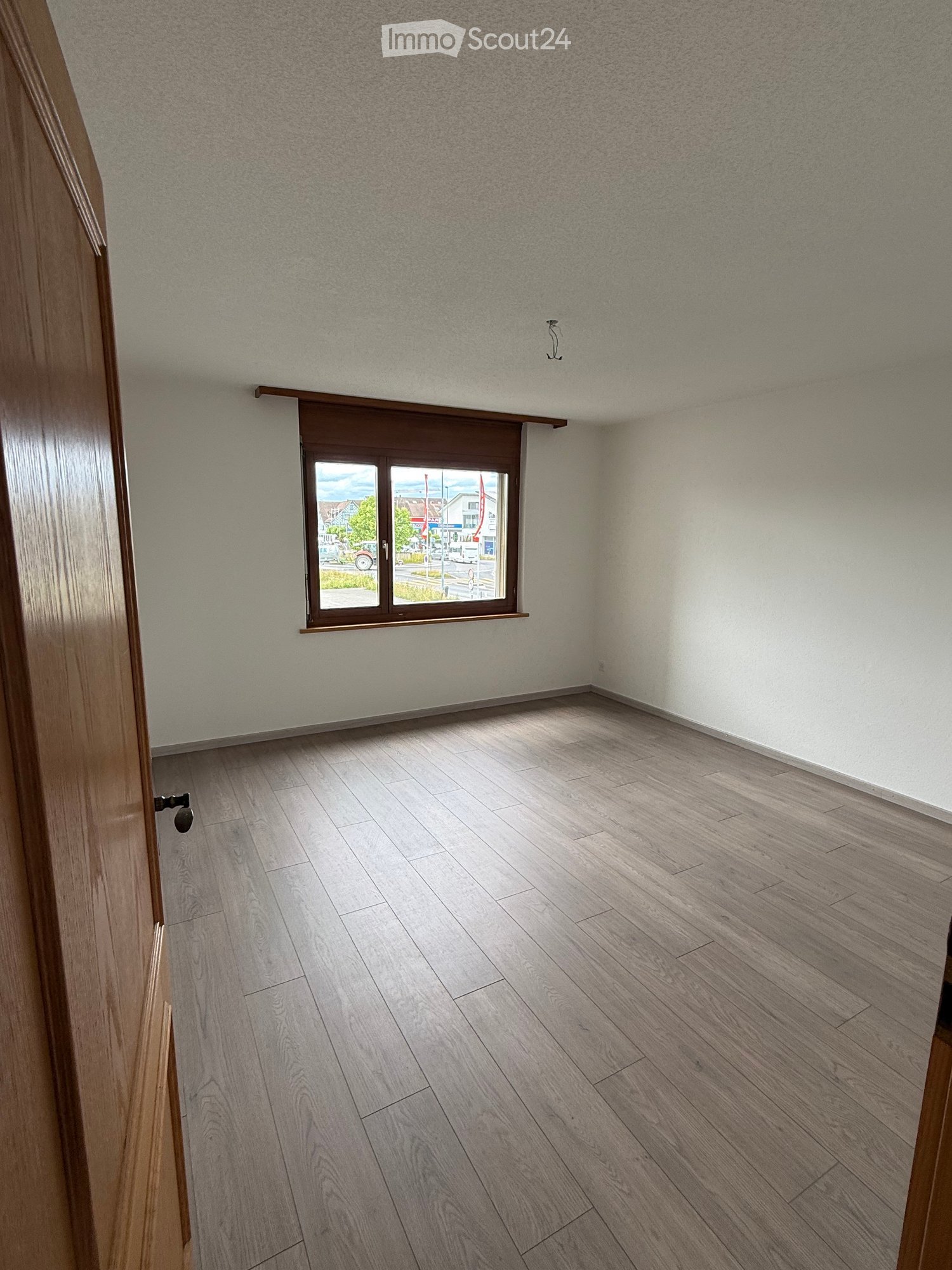 Empty room with light-colored wooden flooring, white walls, and a large window with a brown frame and blinds. The room has a door on the left side.