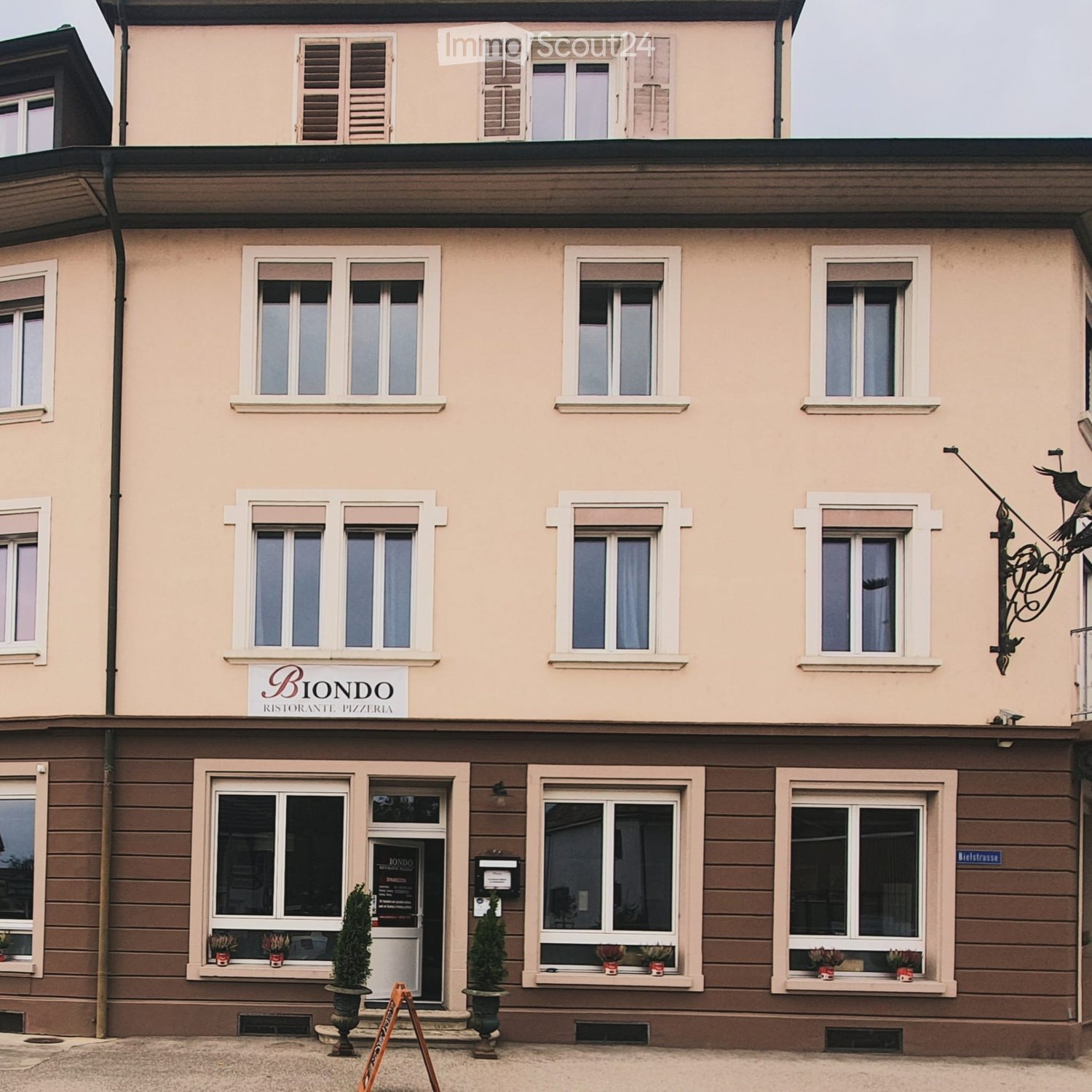 Two-story building with light pink facade, multiple windows, and a sign reading "Biondo Ristorante Pizzeria" above the entrance. The ground floor has a wooden panel facade with potted plants in the windows and a decorative metal bird sculpture on the righ