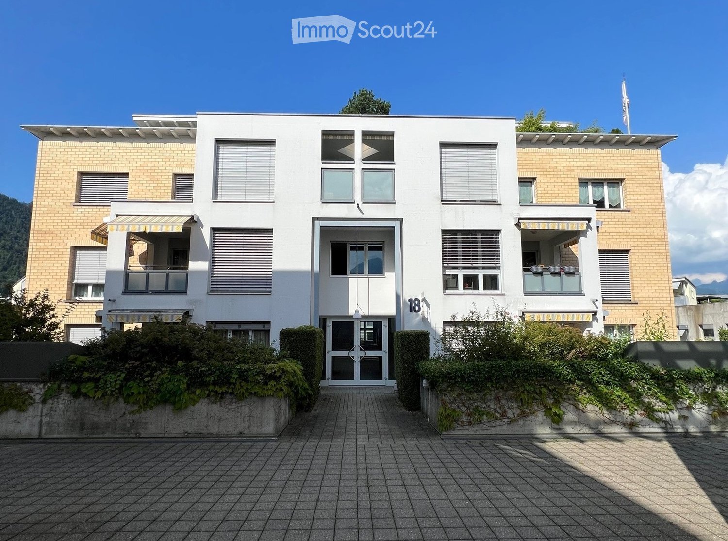 Modern two-story building with white facade, large windows, and balconies. Features a central entrance with glass doors and a small garden area in front.