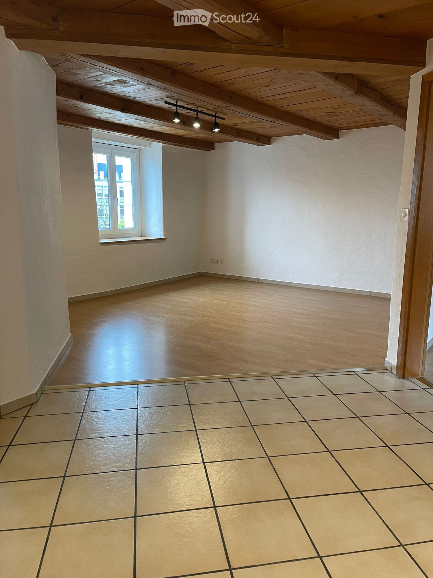 Empty room with wooden floor and exposed wooden ceiling beams, featuring a single window and recessed lighting.