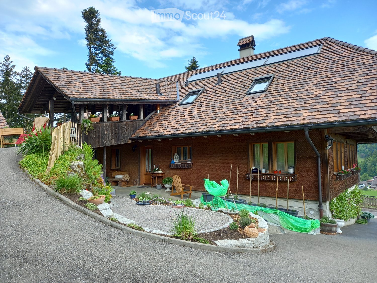 Two-story house with brown tiled roof, multiple windows, and a wooden balcony. The house features a well-maintained garden with potted plants and a wooden chair on the patio.