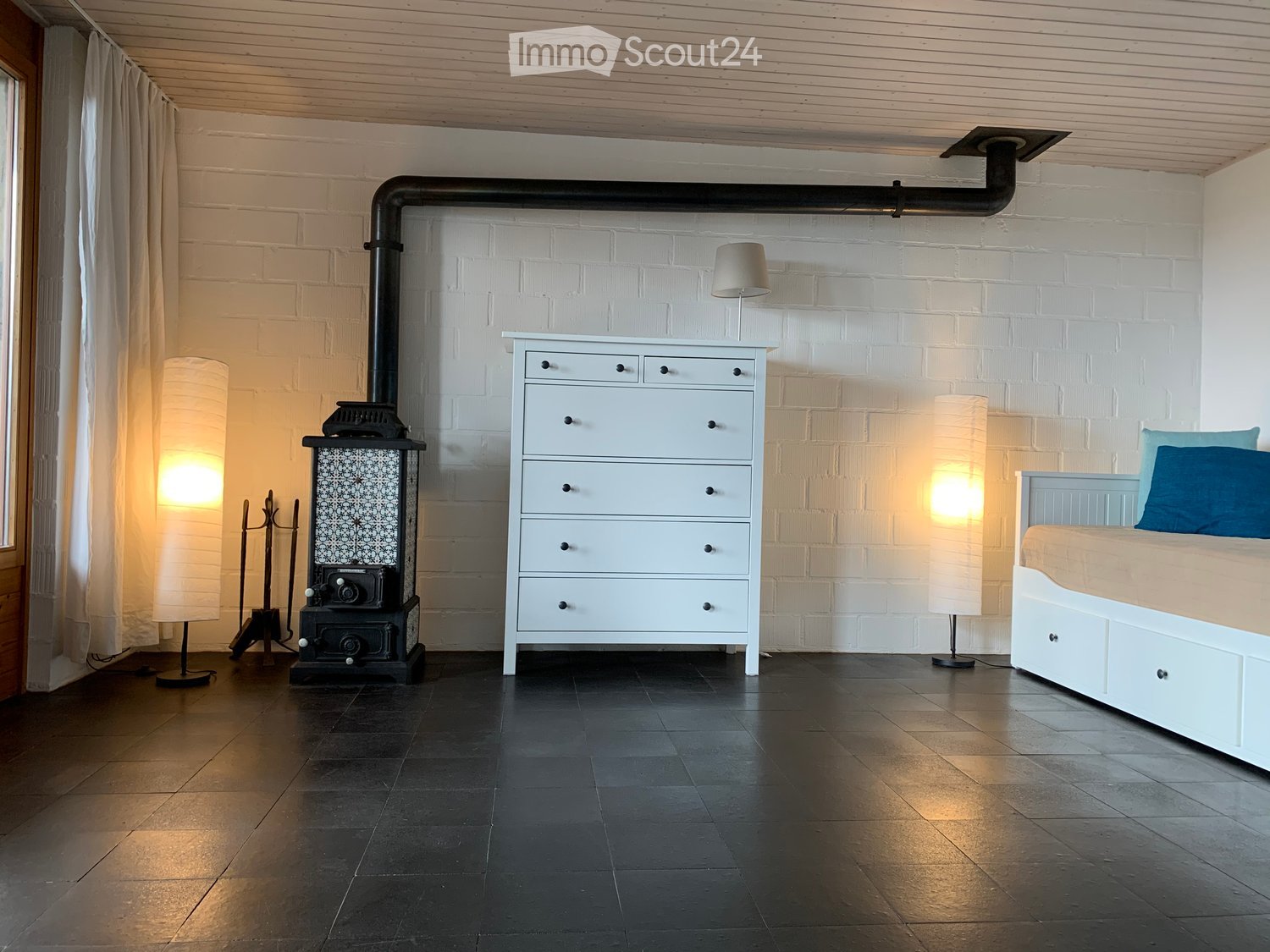 Modern bedroom with a white brick accent wall, a fireplace, a bed with blue pillows, a chest of drawers, and floor lamps.