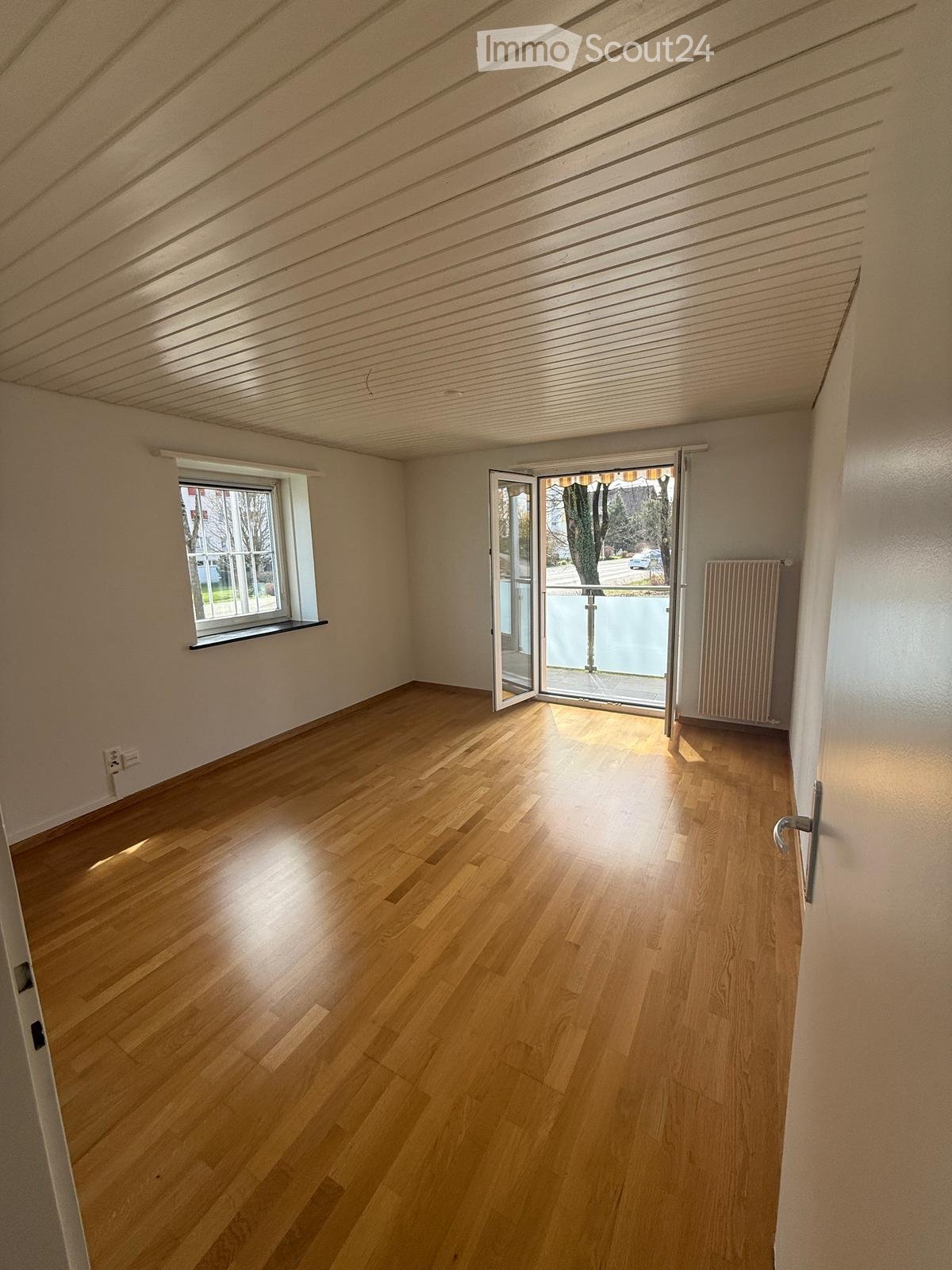 Empty room with wooden flooring, white walls, large window, and sliding glass door leading to a balcony.