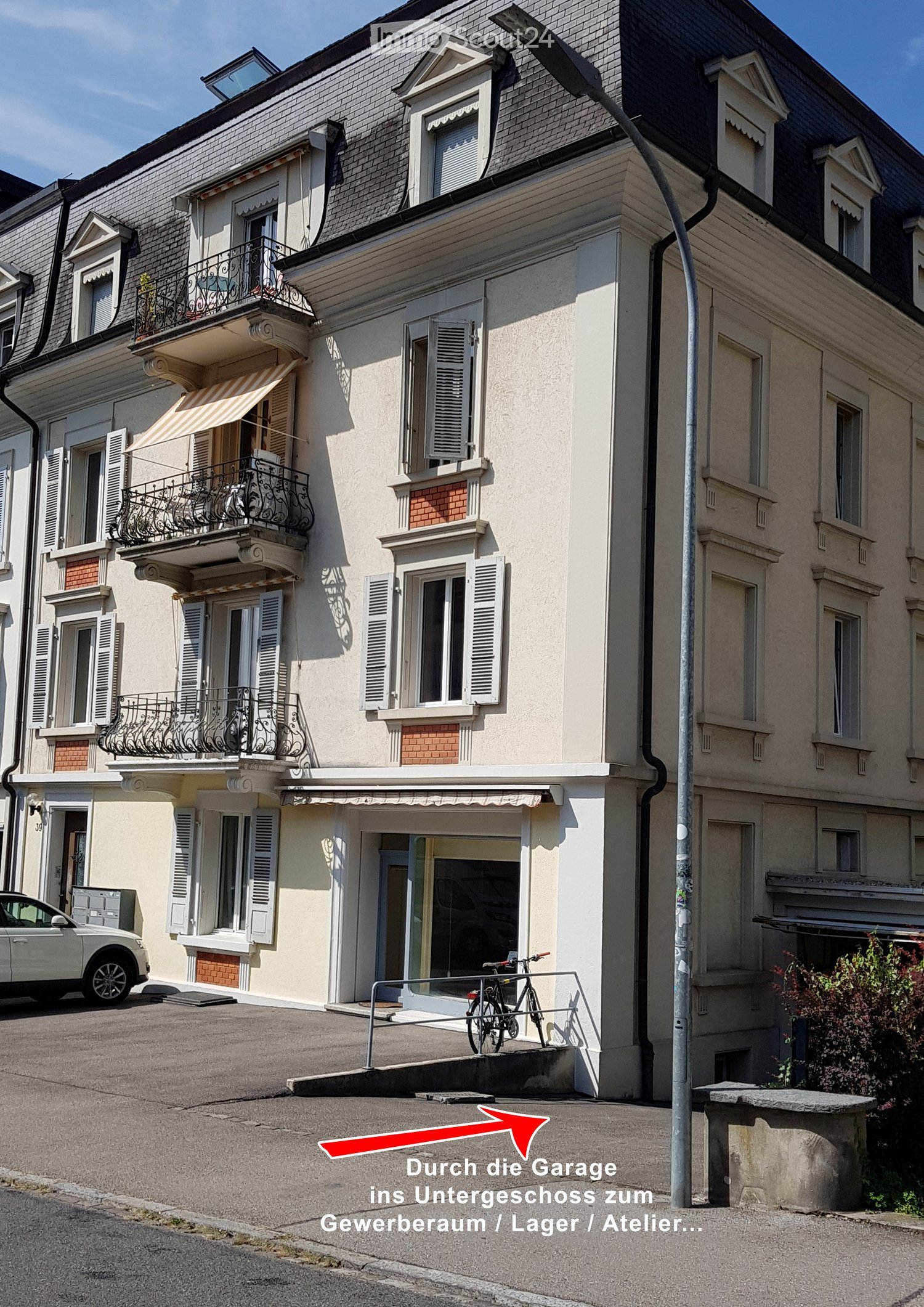 Four-story residential building with balconies, shutters, and a garage entrance. The ground floor has a storefront with glass doors and a bicycle parked nearby.