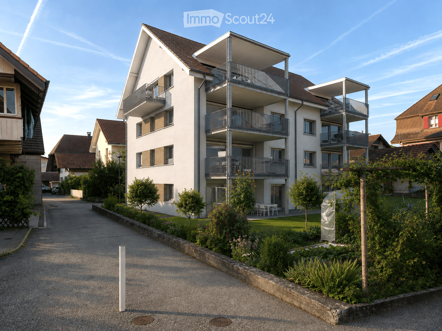 Modern multi-story residential building with balconies, surrounded by greenery and a paved pathway.