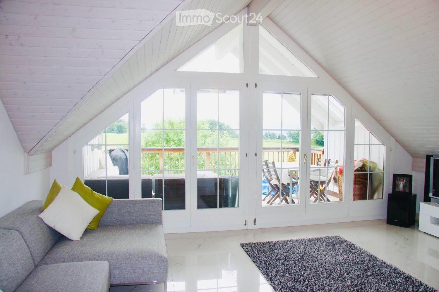 Bright, airy living space with a grey sofa, colorful pillows, a coffee table, and a TV. Large windows and sliding doors open to a patio with outdoor furniture.